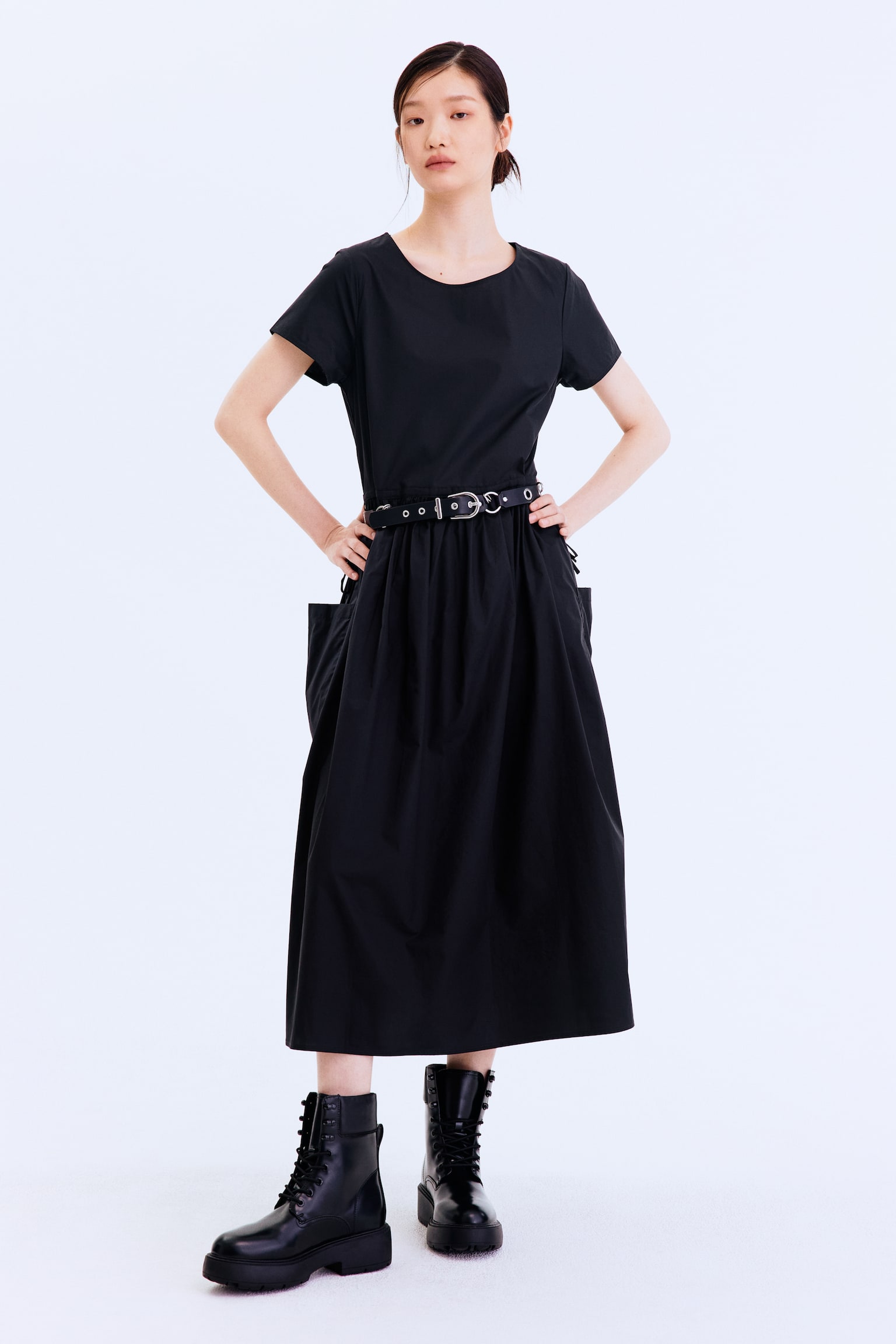 Một chiếc váy đen có cổ tròn, tay ngắn và chân váy midi xòe đầy đủ với một túi vá lớn ở bên trái, được thắt eo bằng một chiếc thắt lưng đen với khóa bạc và lỗ xỏ. - 1