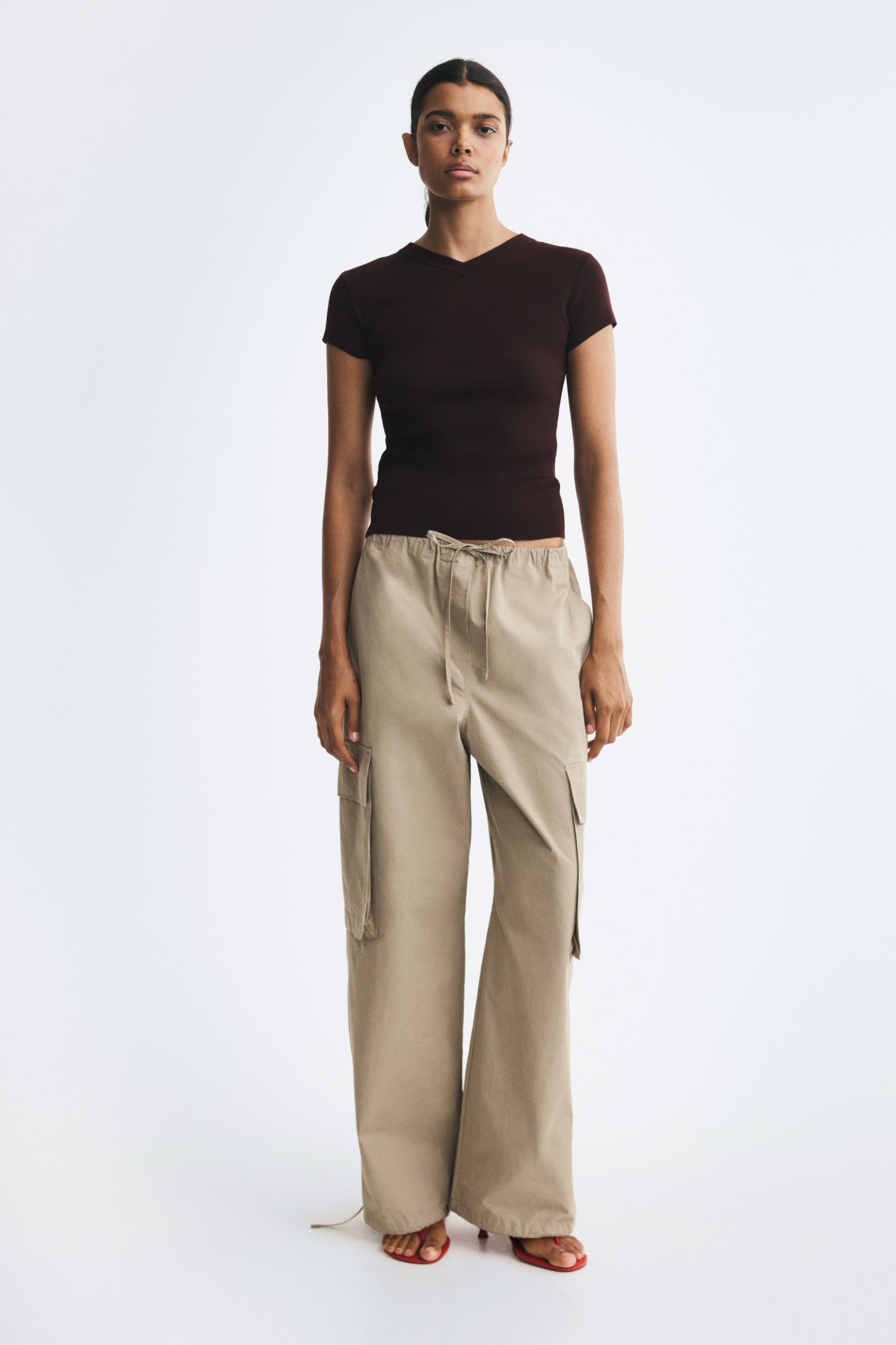 View larger image: A dark brown fitted knit tee featuring short sleeves and a rib-trimmed V-neck is being worn by the model. The tee is styled with wide-leg beige cargo pants.