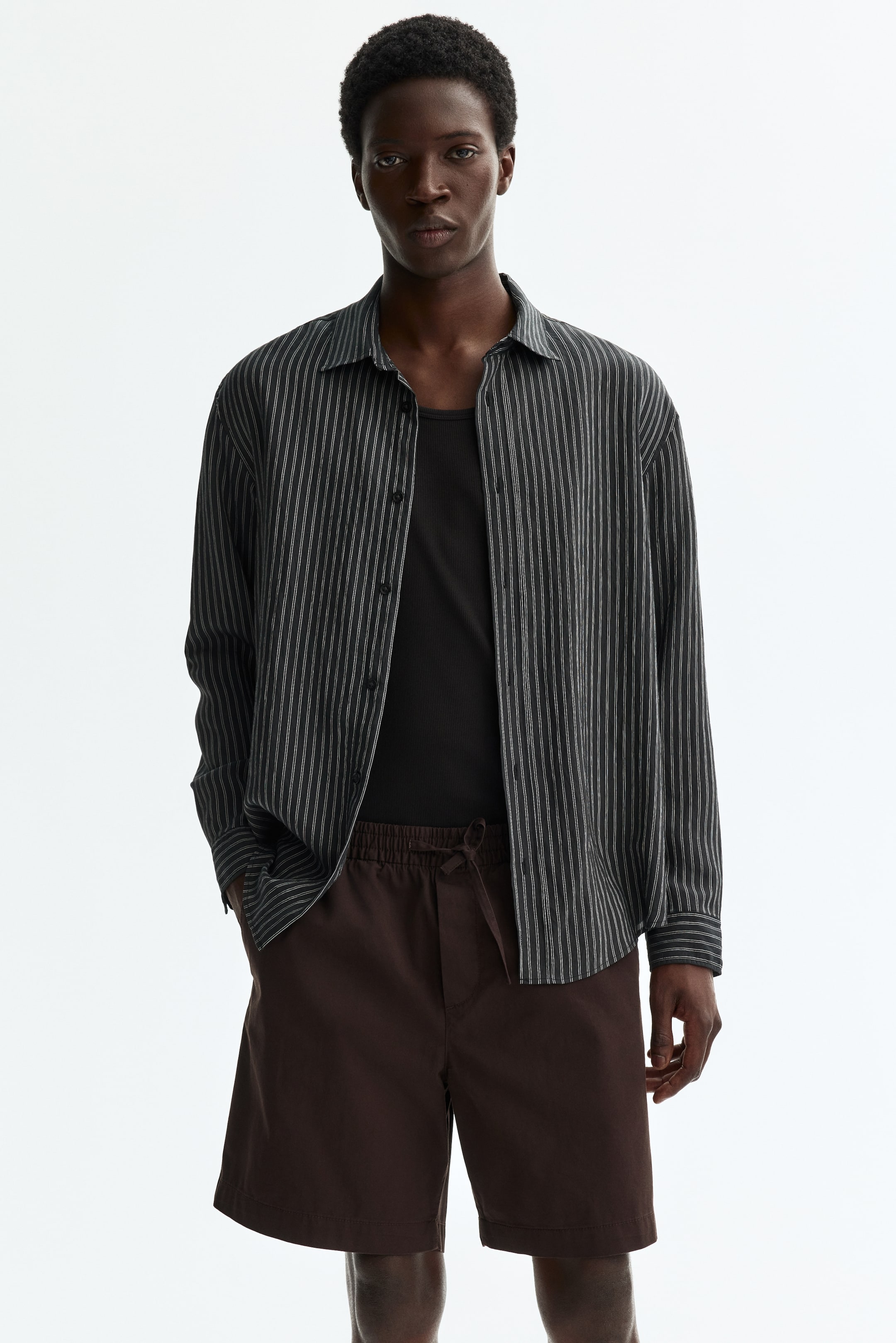View larger image: A gray long-sleeve shirt features thin white vertical stripes and is styled open, revealing a black ribbed tank top underneath. It showcases a pointed collar and a front button closure.