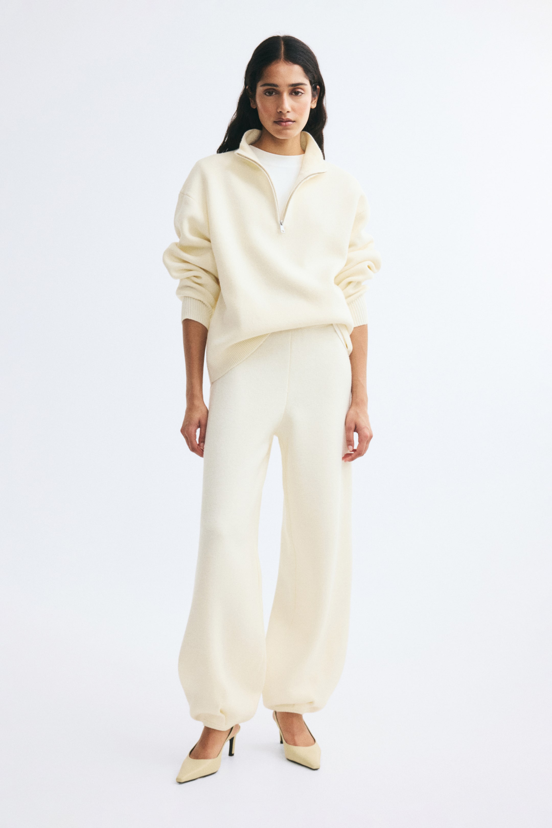 Agrandir l'image: Ce pantalon finement tricoté couleur crème est présenté de face, avec une coupe taille haute et une silhouette ballon volumineuse. Les jambes se resserrent jusqu’à des poignets côtelés et plissés aux chevilles, et le pantalon est associé à un pull crème assorti à fermeture éclair quart de longueur ainsi qu’à des escarpins beige.