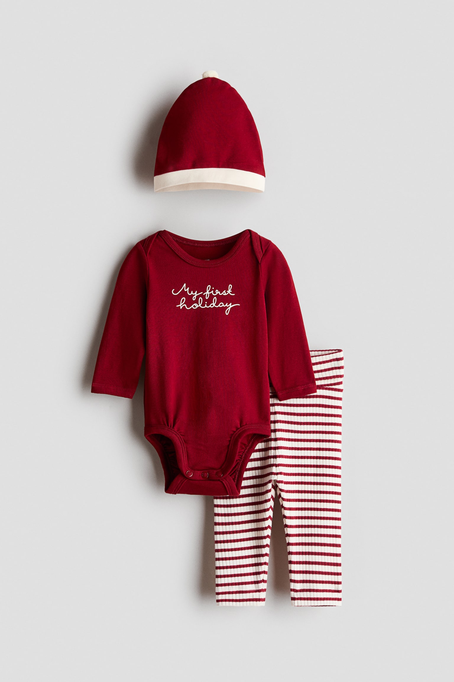 Un set per neonato in tre pezzi Ã¨ mostrato frontalmente, composto da un body a maniche lunghe color rosso scuro con la scritta "My first holiday" in corsivo bianco sul petto. Il set include pantaloni a coste con righe orizzontali rosse e bianche e un cappellino rosso abbinato con risvolto bianco ripiegato e un piccolo pon-pon sulla sommitÃ .