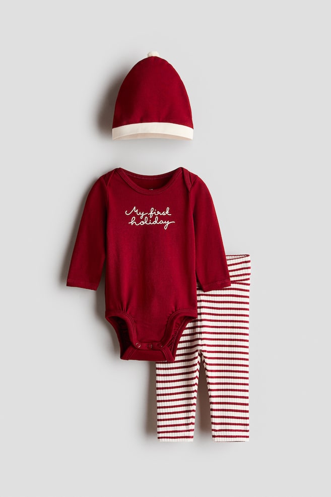 A three-piece baby set is shown from the front, featuring a long-sleeved dark red bodysuit with "My first holiday" written in white cursive script across the chest. The set includes red and white horizontally striped ribbed pants and a matching red hat with a white folded brim and a small pom-pom on top. - 1