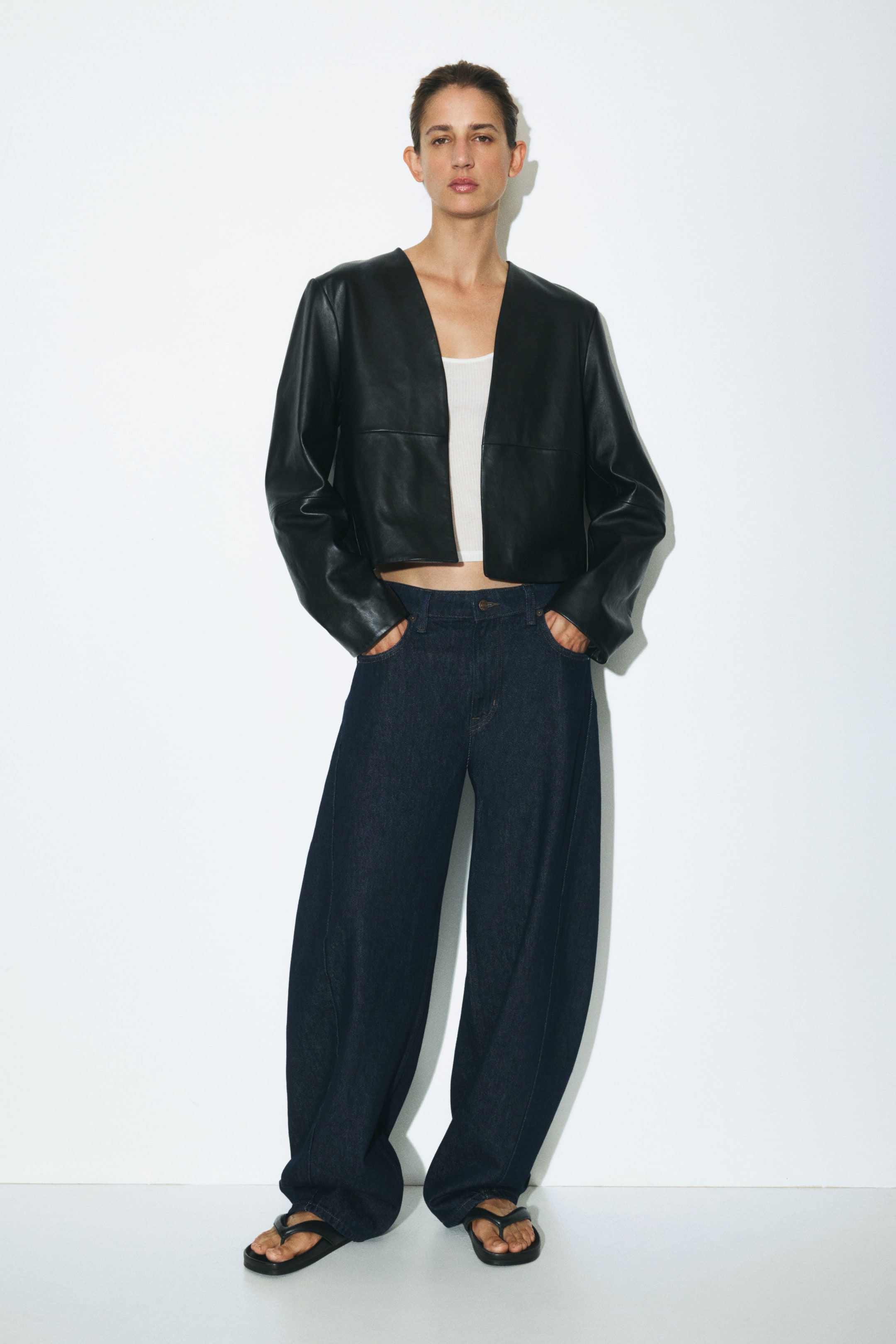 Visa större bild: Mörkblå jeans i denim med normal midja och böjda, voluminösa ben bärs av modellen, som står med händerna i framfickorna. Jeansen stylas med en kort svart skinnjacka utan krage, en vit topp och svarta tongsandaler.