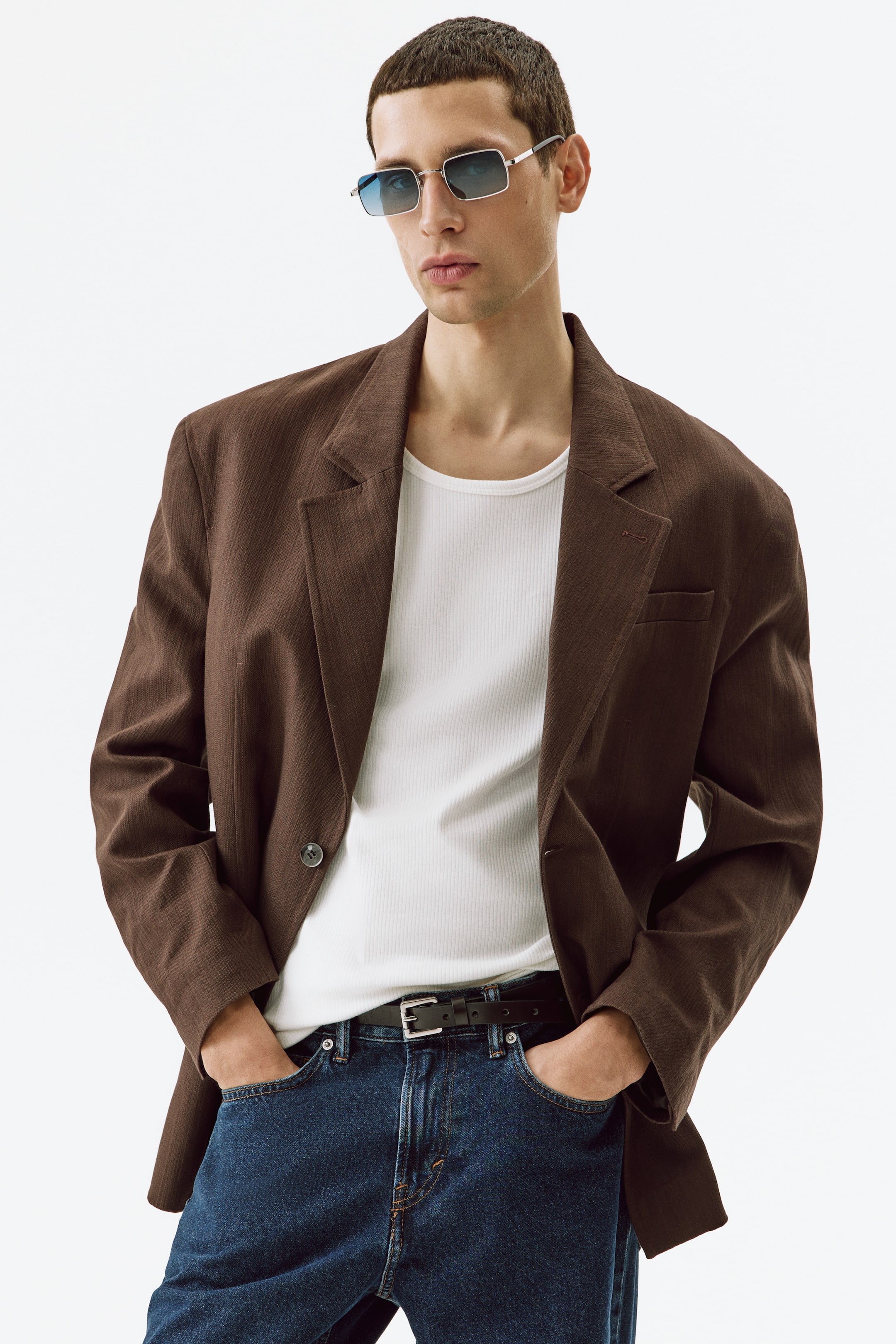 Visa större bild: En brun blazer presenteras, med kavajslag, en subtil vertikal textur och enknappsstängning. Den bärs öppen över ett vitt ribbat linne, mörkblå jeans och ett svart bälte, vilket visar upp dess avslappnade passform från en lätt höger vinkel.