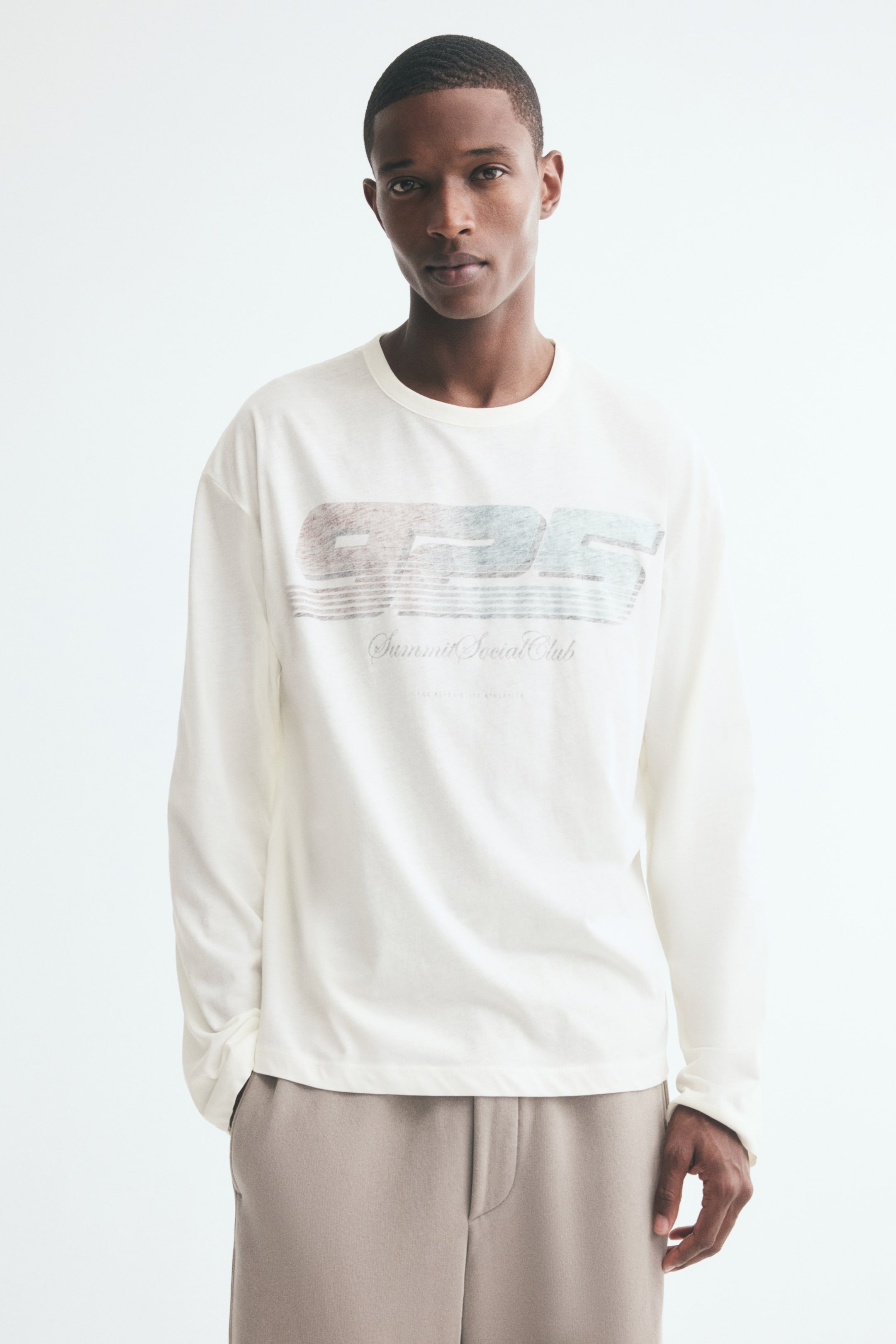View larger image: An off-white, long-sleeve tee featuring a graphic print on the front, a round neckline, and dropped shoulders is being worn by the model. The relaxed-fit, boxy tee is styled with beige trousers.