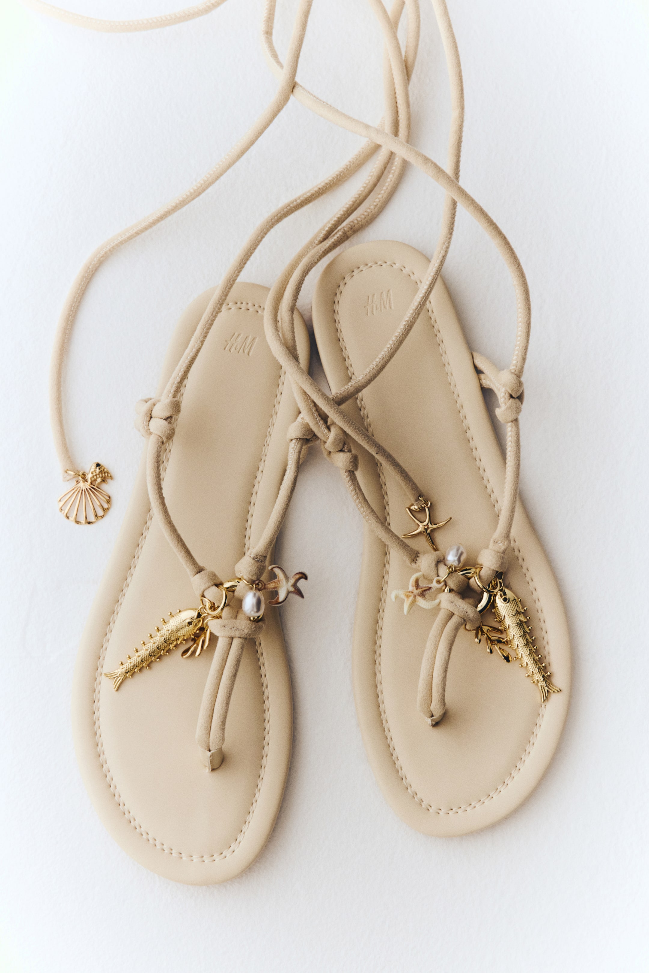 Agrandir l'image: Une paire de sandales plates beige est présentée, avec leurs longues lanières qui se croisent et s'étendent vers le haut. Chaque sandale est dotée d'une semelle plate avec un logo H&M embossé et est ornée de breloques dorées en forme de poisson, d'étoile de mer et de perles, tandis qu'une breloque supplémentaire en forme de coquillage doré est visible sur l'une des lanières prolongées.