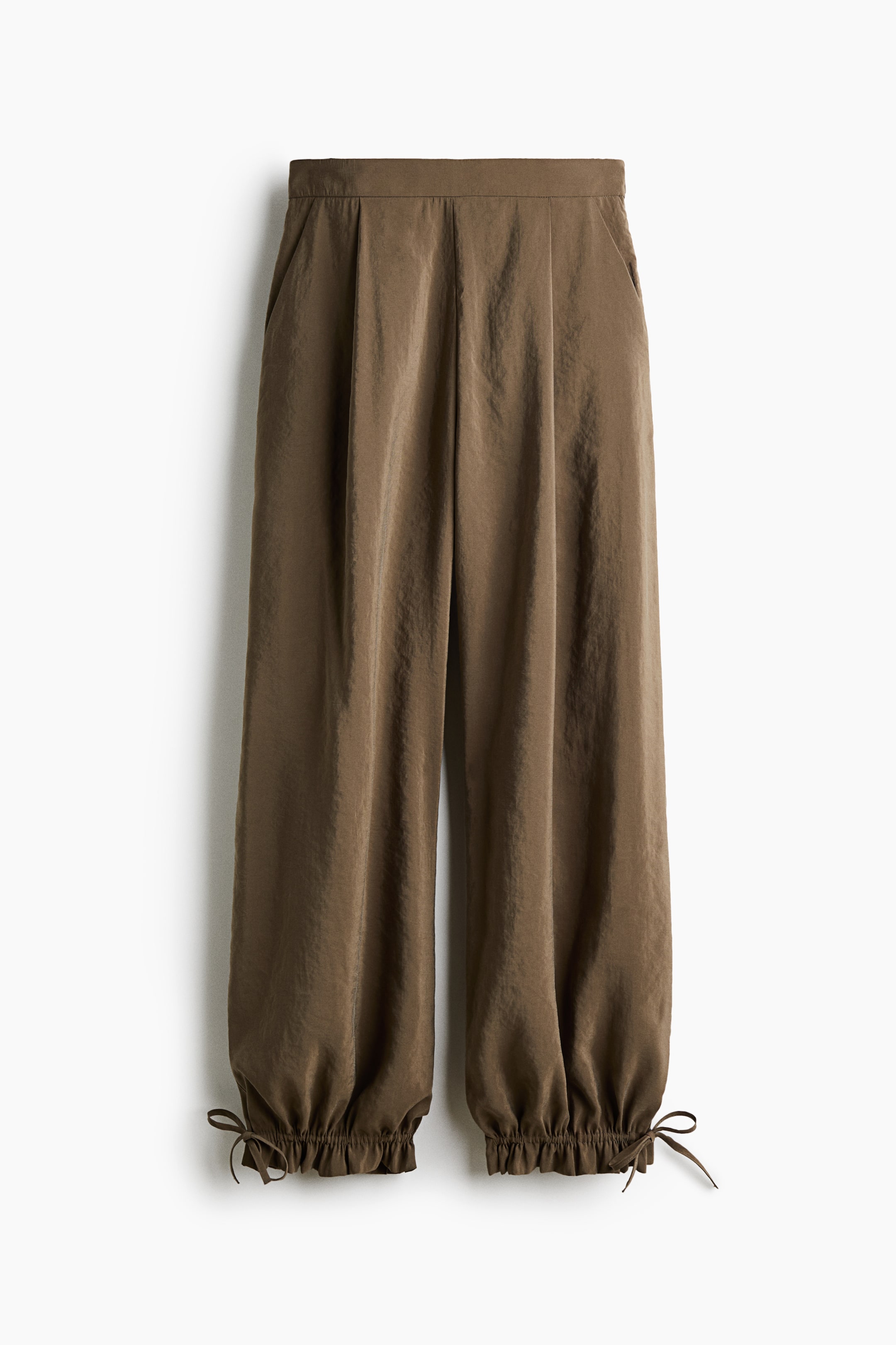 View larger image: This pair of brown, wide-leg trousers is shown from the front, featuring a high-waisted waistband with front pleats and side pockets. The loose-fitting legs drape into gathered cuffs secured with adjustable drawstring bows at the outer ankles.