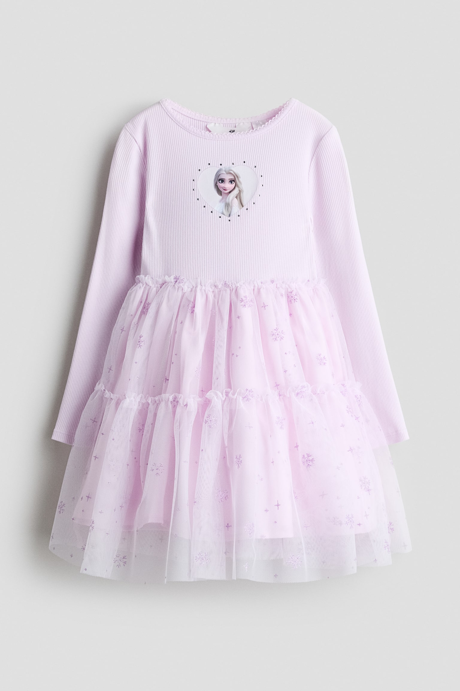 A light pink long-sleeved dress is shown from the front, featuring a ribbed knit bodice with a heart-shaped graphic of Elsa on the chest. The dress features a tiered, flared tulle skirt decorated with glittery snowflake and star motifs.