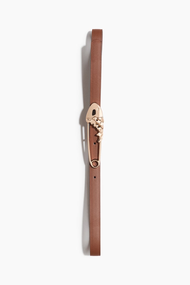 A narrow brown belt is featuring a gold-toned metal pin buckle, which is shaped like a safety pin with an intricate fish head and scaled body design. - 1