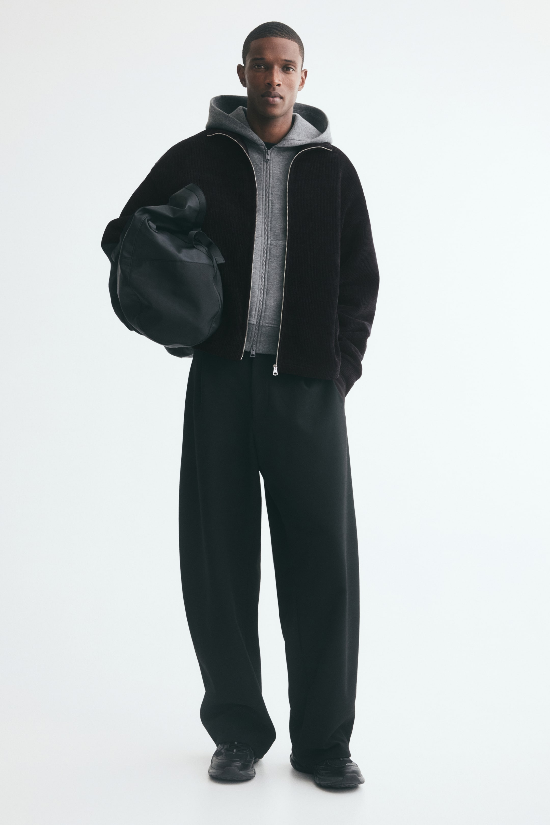 View larger image: A dark brown chenille track jacket featuring a relaxed fit, dropped shoulders, and a stand collar is being worn unzipped by the model over a grey zip-up hoodie. The jacket is styled with black wide-leg trousers, and the model is carrying a black bag under one arm.
