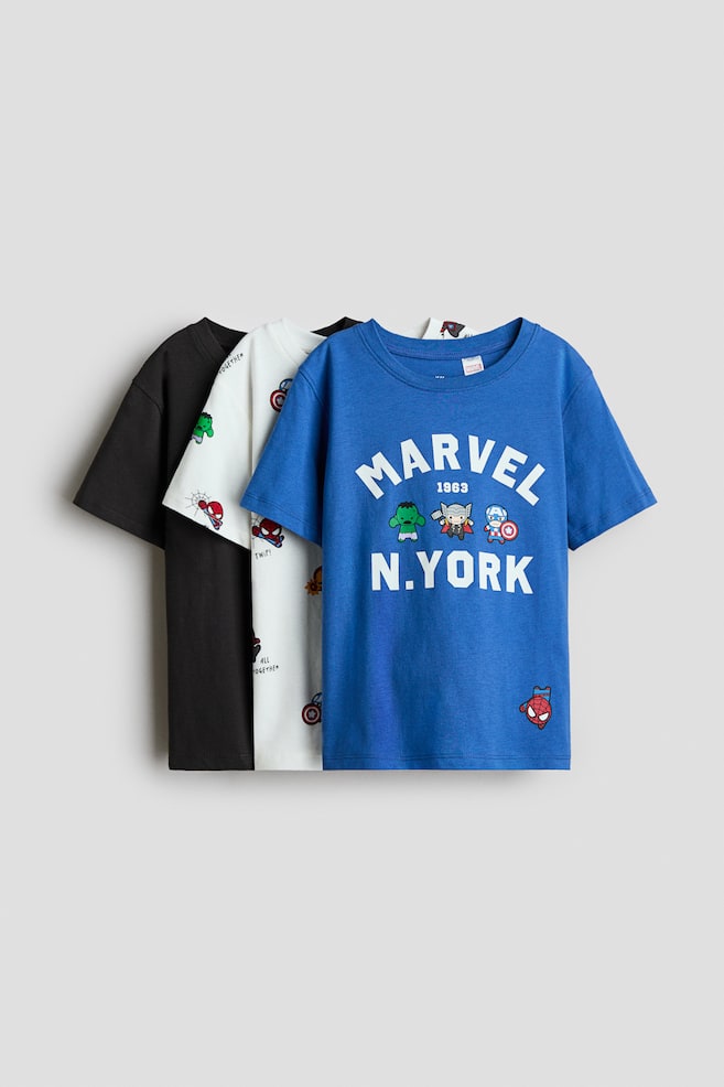 Three classic cotton-jersey t-shirts are shown facing front in a layered arrangement, displaying their short sleeves and rib-trimmed crew necklines. The front blue t-shirt is featuring a printed white "MARVEL 1963 N.YORK" graphic with small cartoon characters, while the middle white t-shirt is displaying an all-over superhero print, and the back t-shirt is solid black. - 1