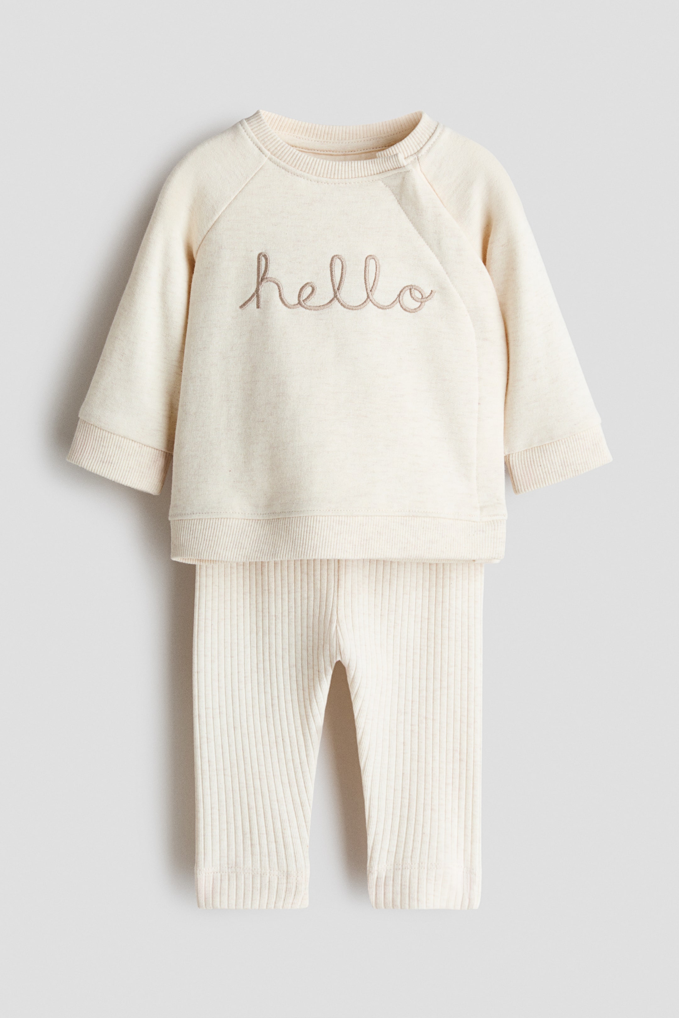 Größeres Bild anzeigen: Ein cremefarbenes Baby-Set wird von vorne präsentiert, bestehend aus einem leichten Sweatshirt-Oberteil mit Rundhalsausschnitt, langen Raglanärmeln und "hello" in Schreibschrift auf der Brust gestickt. Das Oberteil zeigt kleine Knöpfe an der rechten Schulternaht und ist mit gerippten Bündchen und Saum versehen, während passende gerippte Jersey-Leggings das Set mit einem breiten elastischen Bund und gerippten Säumen vervollständigen.