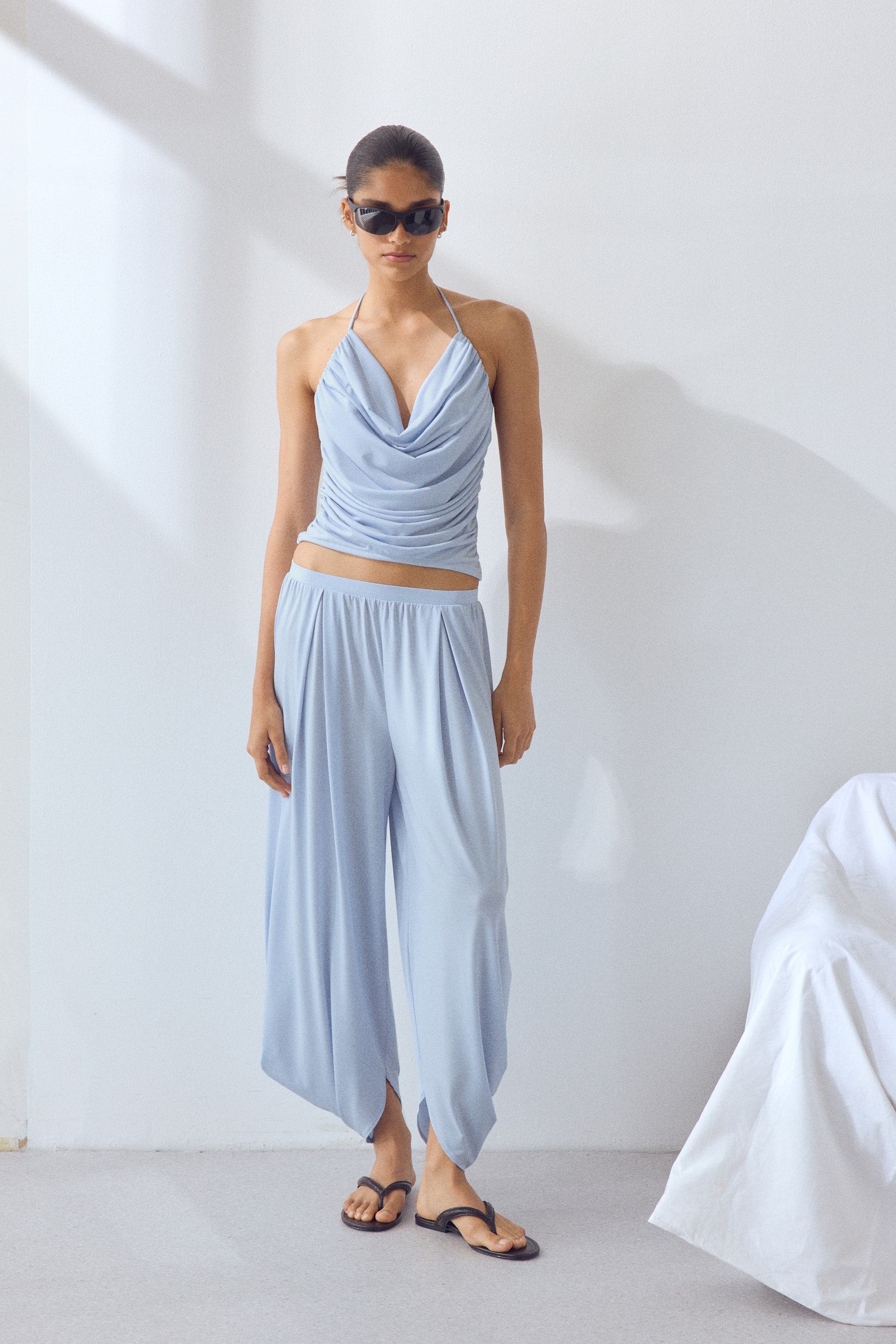 View larger image: The model is donning draped pull-on trousers in a soothing light blue hue. These trousers boast an elasticated waistband, a wide-leg silhouette, and a tapered ankle adorned with a split hem. Paired elegantly with a coordinating draped halter top, the ensemble exudes a chic and contemporary vibe. The model is posed standing.