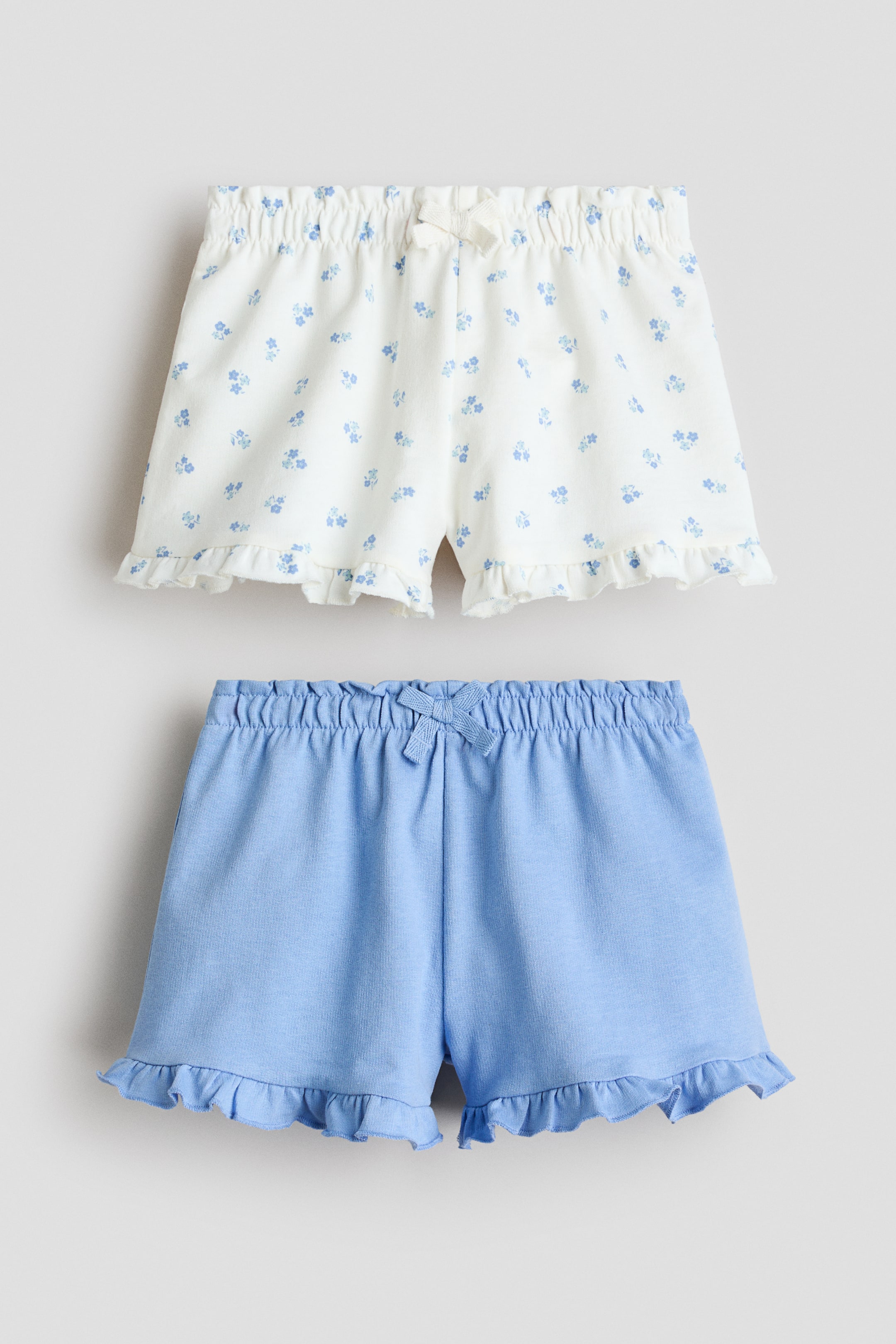 Vis større bilde: To par shorts blir presentert forfra, begge med et lett sweatstoff, en elastisk, volangkantet midje med en liten dekorativ sløyfe, og volangkantede falder. Det ene paret viser et hvitt stoff med et blått blomstertrykk, og det andre viser et ensfarget lyseblått stoff.