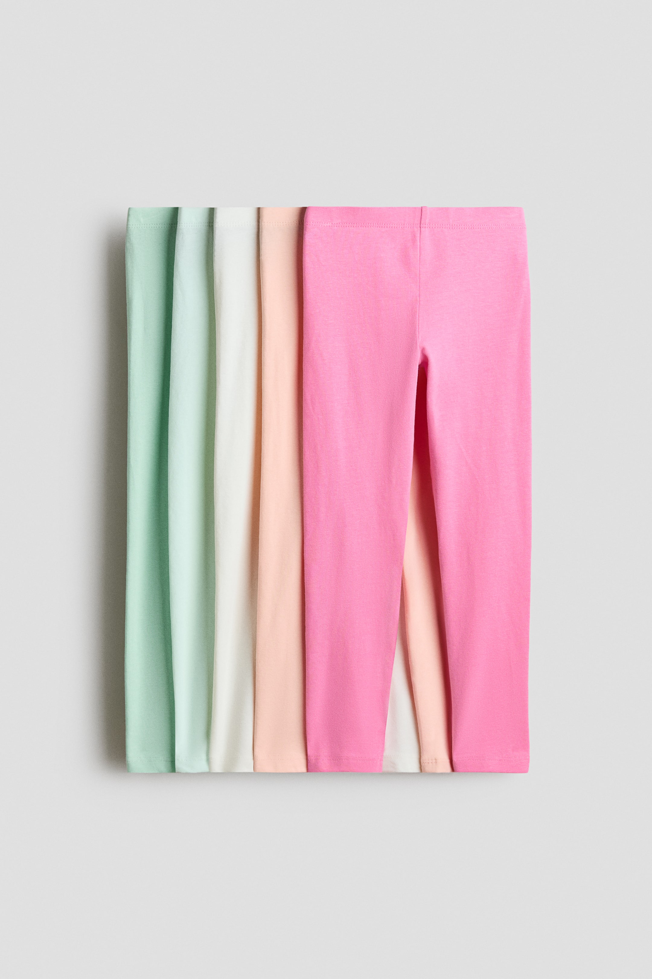 View larger image: A set of five soft cotton jersey leggings with elastic waistbands is shown stacked horizontally in shades of mint green, off-white, light peach, bright pink, and pale cream, with the bright pink pair facing forward to reveal the elastic waistband and upper leg shape.