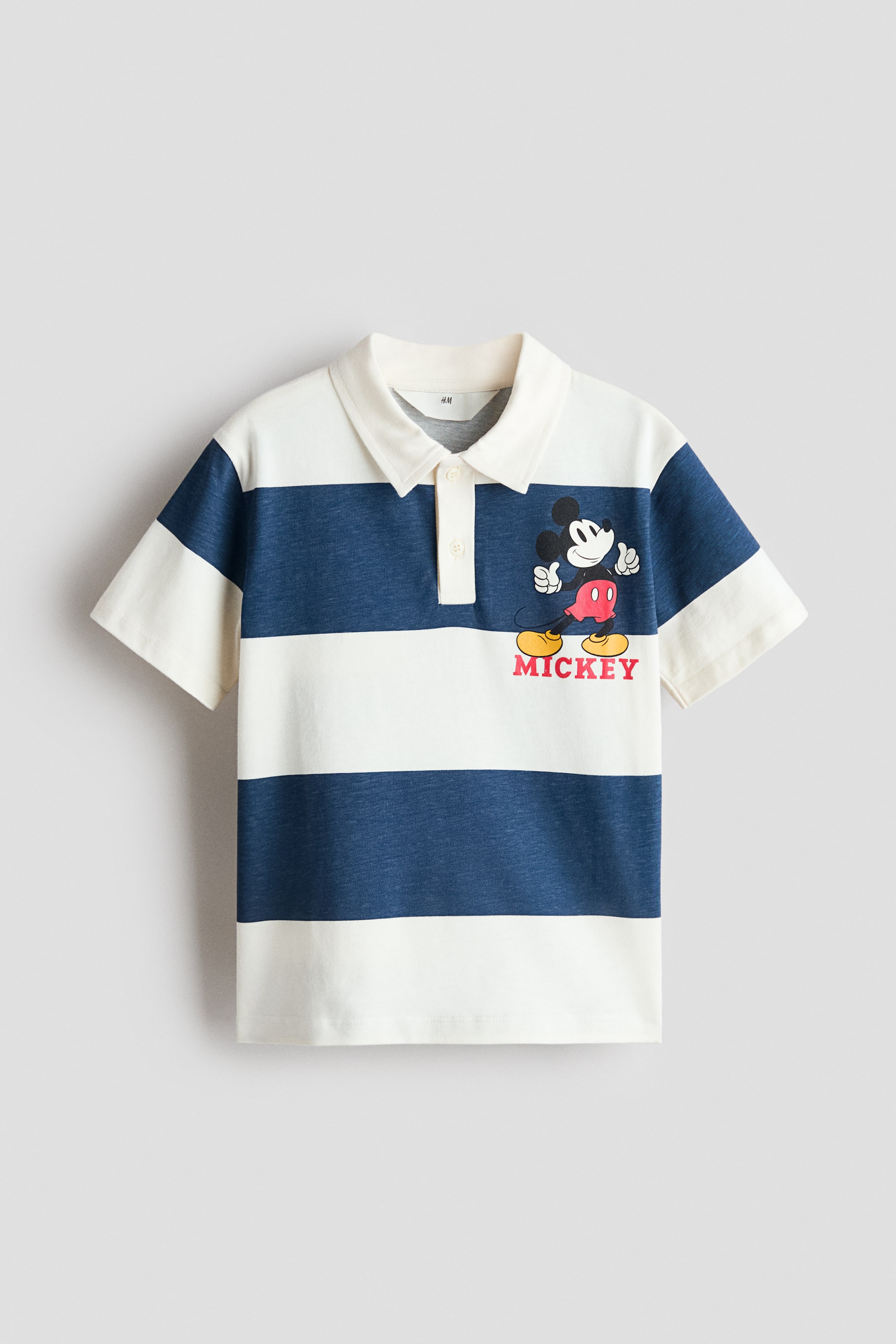Grösseres Bild ansehen: Ein Poloshirt wird von vorne präsentiert, mit weißen und blauen horizontalen Streifen. Es ist mit einem weißen Kragen, einer Zweiknopfleiste und kurzen Ärmeln gestaltet und zeigt ein Mickey Mouse-Motiv sowie den Schriftzug „MICKEY“ auf der linken Brust.