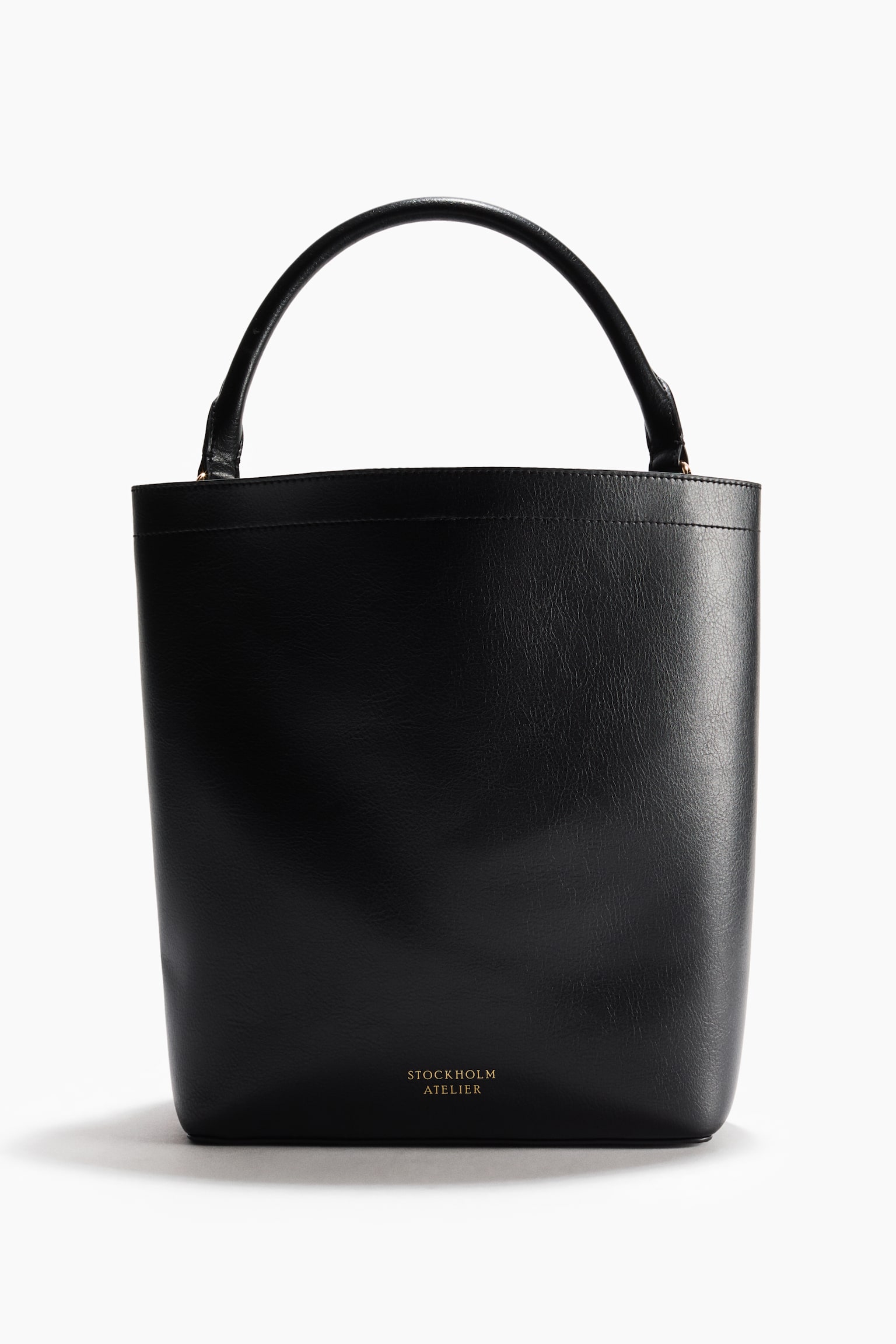 Um saco de balde preto está voltado para a frente, exibindo uma textura suave, uma única alça superior curva e o logótipo 'STOCKHOLM ATELIER' em dourado na parte inferior da frente. Apresenta um fecho magnético oculto na parte superior, uma alça de ombro destacável e ajustável, e um divisor interno que cria dois compartimentos, um dos quais é maior que o outro, criando um efeito desleixado; também é forrado. - 1