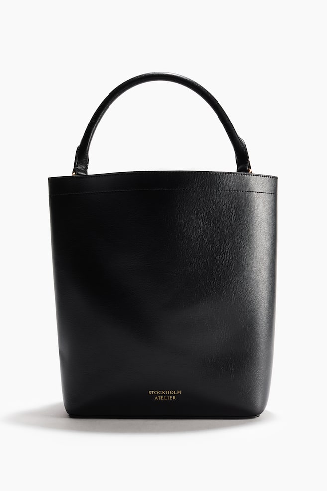 Un sac seau noir est orienté vers l'avant, affichant une texture lisse, une seule poignée supérieure incurvée, et le logo 'STOCKHOLM ATELIER' en or sur sa partie inférieure avant. Il est doté d'une fermeture magnétique dissimulée sur le dessus, d'une bandoulière amovible et réglable, et d'un séparateur interne créant deux compartiments, dont l'un est plus grand que l'autre, créant ainsi un effet souple; il est également doublé. - 1