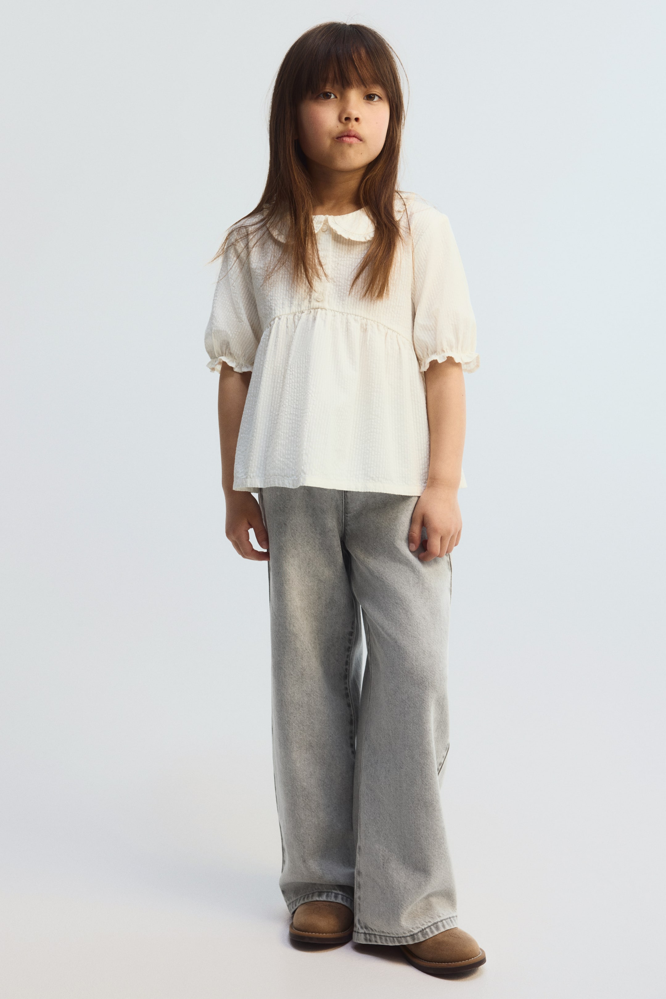 Vis større bilde: En off-white teksturert topp med Peter Pan-krage, frontknapper og korte, rynkede ermer bæres, og gir en avslappet passform. Den er stylet med lysegrå denim bukser med vide ben og brune mokkasiner i semsket skinn, alle vendt fremover.