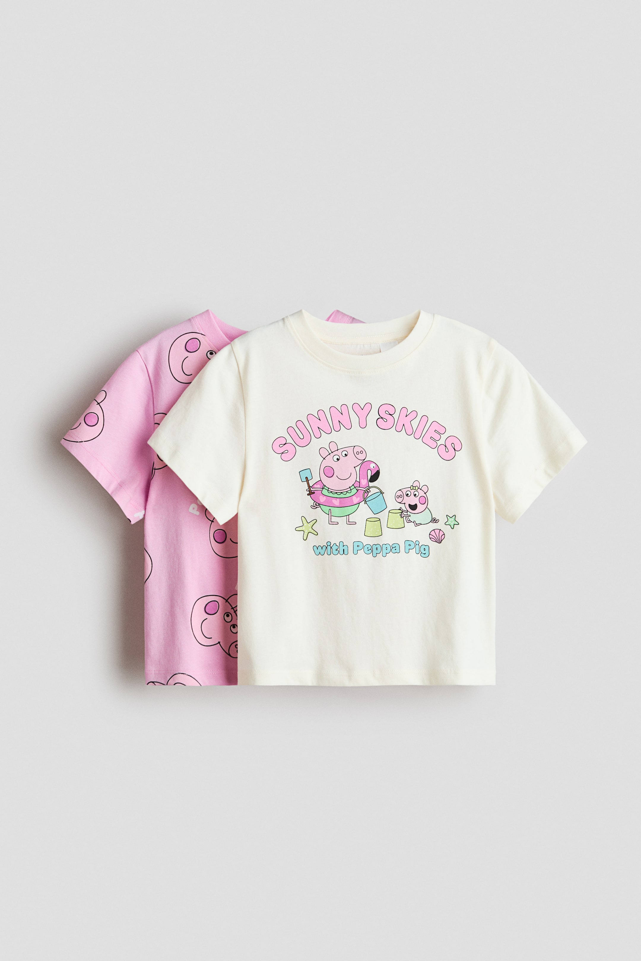 View larger image: A cream short-sleeved t-shirt showcases its front, adorned with a vibrant graphic print of "SUNNY SKIES with Peppa Pig," depicting Peppa and George Pig enjoying a day at the beach. Behind it, a pink short-sleeved t-shirt is layered, featuring an all-over outline print of Peppa Pig's face.