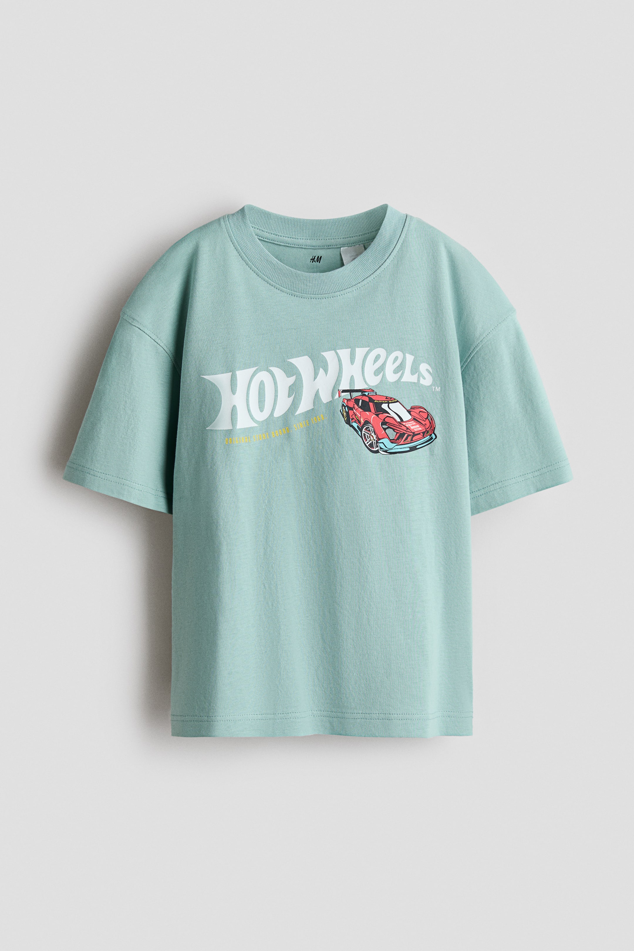 Visualizza l'immagine ingrandita: Una T-shirt oversize in jersey di cotone stampato color teal chiaro presenta il suo davanti, con il logo bianco Hot Wheels e una grafica di un'auto da corsa rossa. È progettata con un girocollo a coste e spalle scese.
