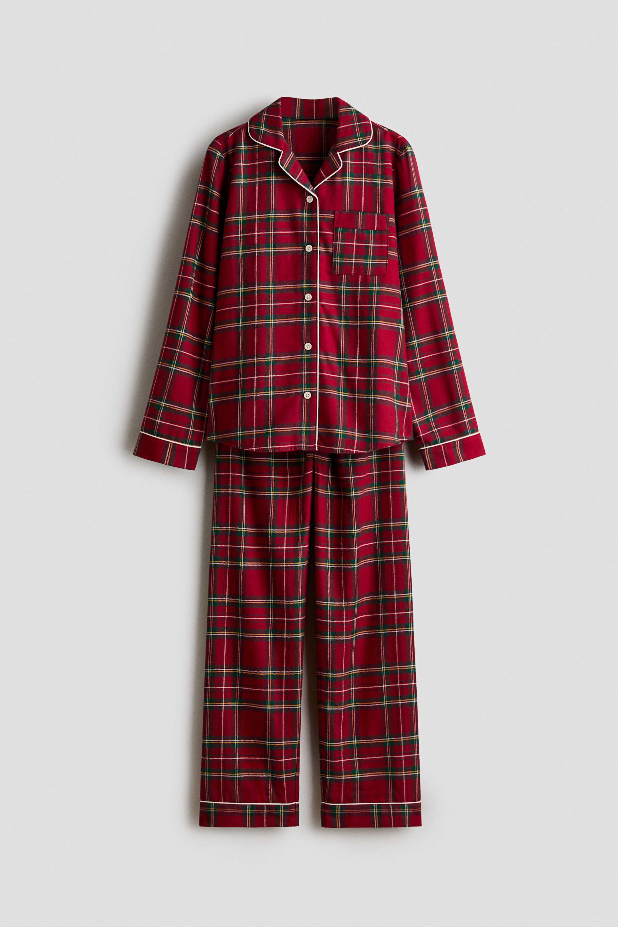 Agrandir l'image: Un ensemble de pyjama deux pièces à motif tartan rouge est suspendu de face, mettant en valeur une chemise boutonnée à manches longues et un pantalon droit assorti. La chemise est agrémentée d’un passepoil blanc le long des revers crantés, de la patte de boutonnage et des poignets, avec une seule poche plaquée sur la poitrine gauche.