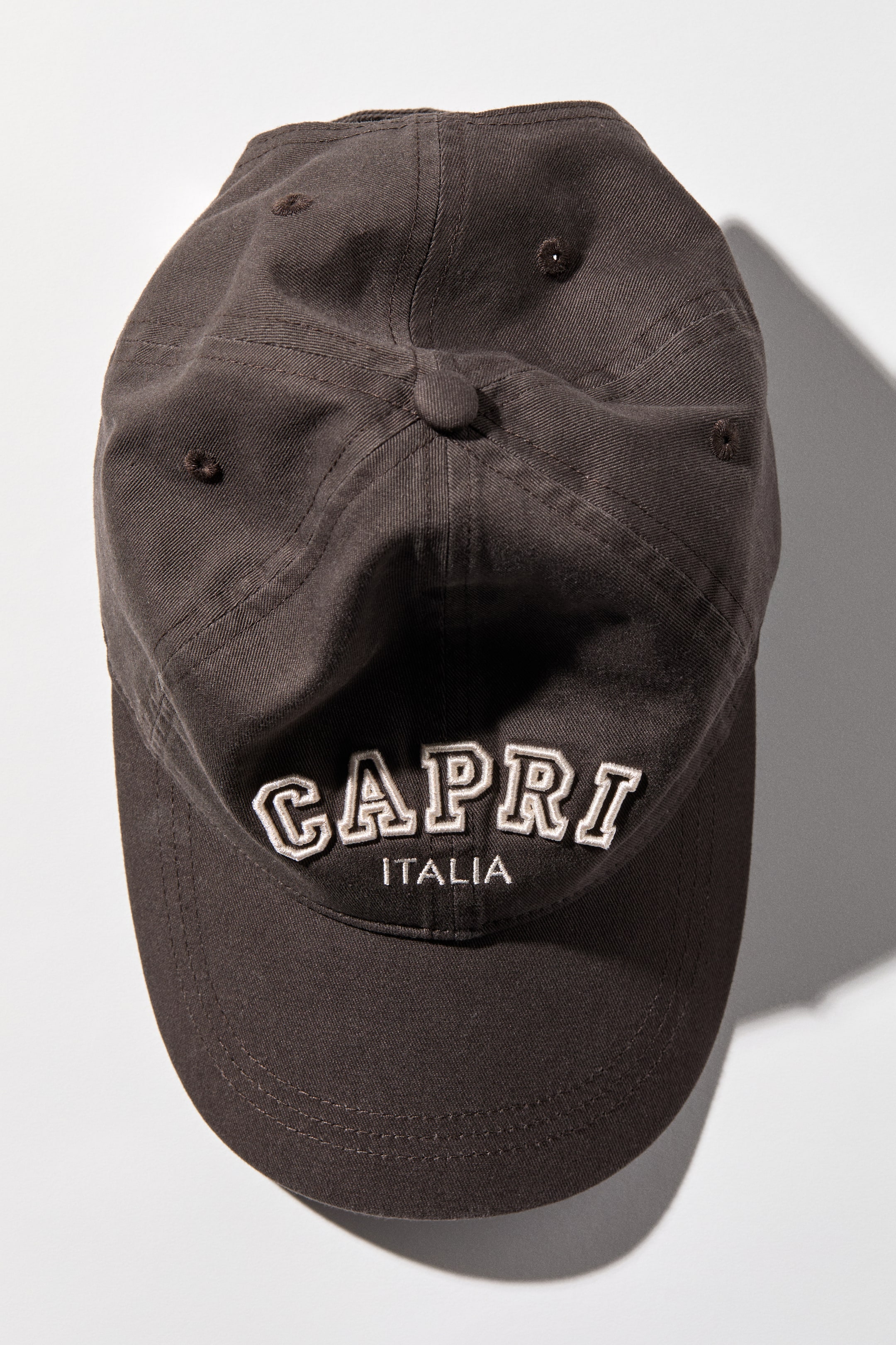 View larger image: A dark brown baseball cap is lying flat, showcasing its top and front, featuring a six-panel construction with ventilation eyelets and a top button. The front panel displays embroidered 'CAPRI' in large, white, outlined block letters with 'ITALIA' in smaller white text beneath it, above a curved brim.