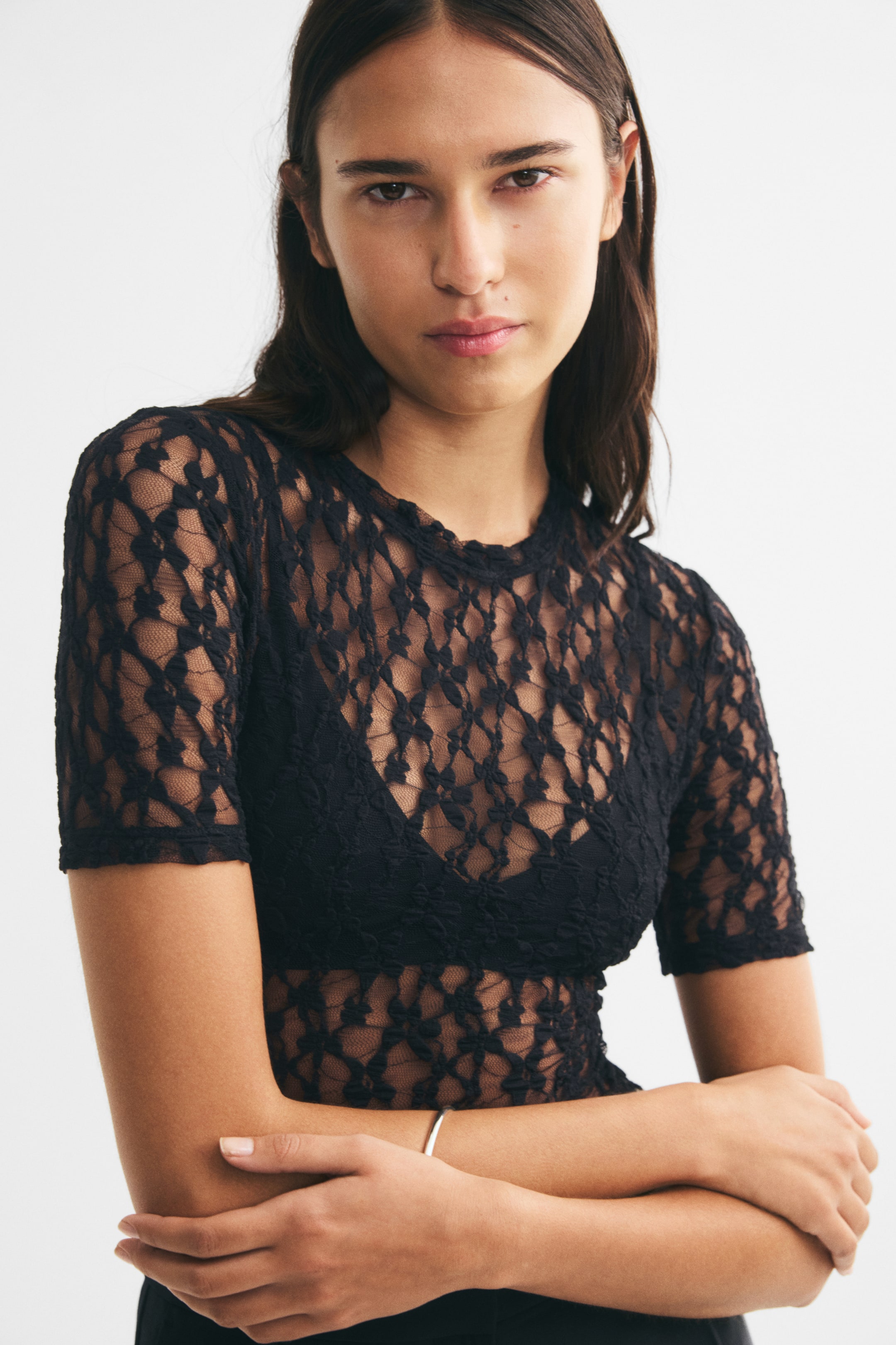View larger image: A black lace bodysuit with short sleeves and a crew neckline is worn by the model over a solid black bra. The sheer lace fabric covers the torso and arms, and the model is posing with her arms crossed.