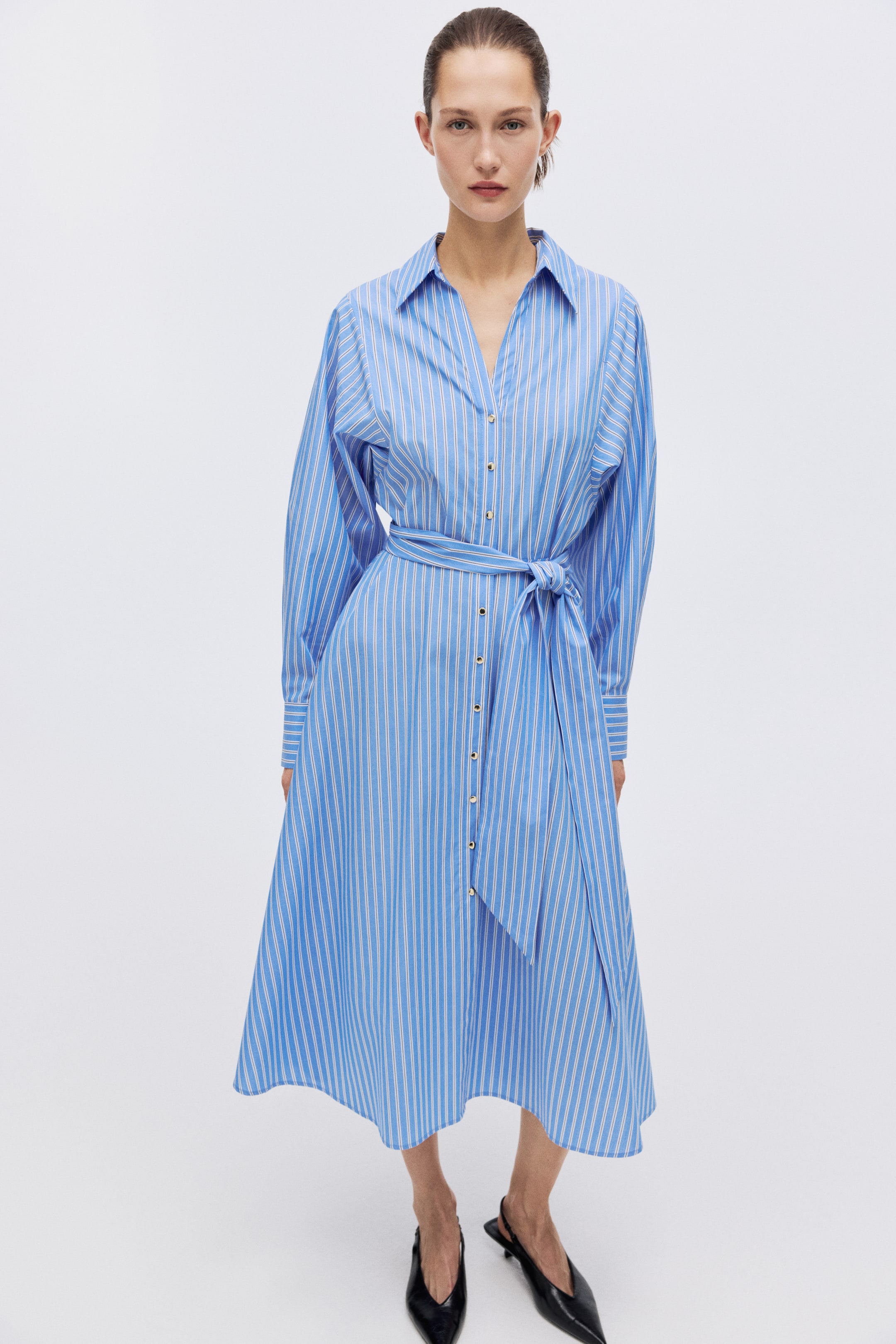 View larger image: A light blue-and-white striped shirt dress features a collared neckline, long sleeves, and a button-front closure with gold-toned buttons. A matching tie belt cinches the waist, and the A-line skirt flows to a midi length.