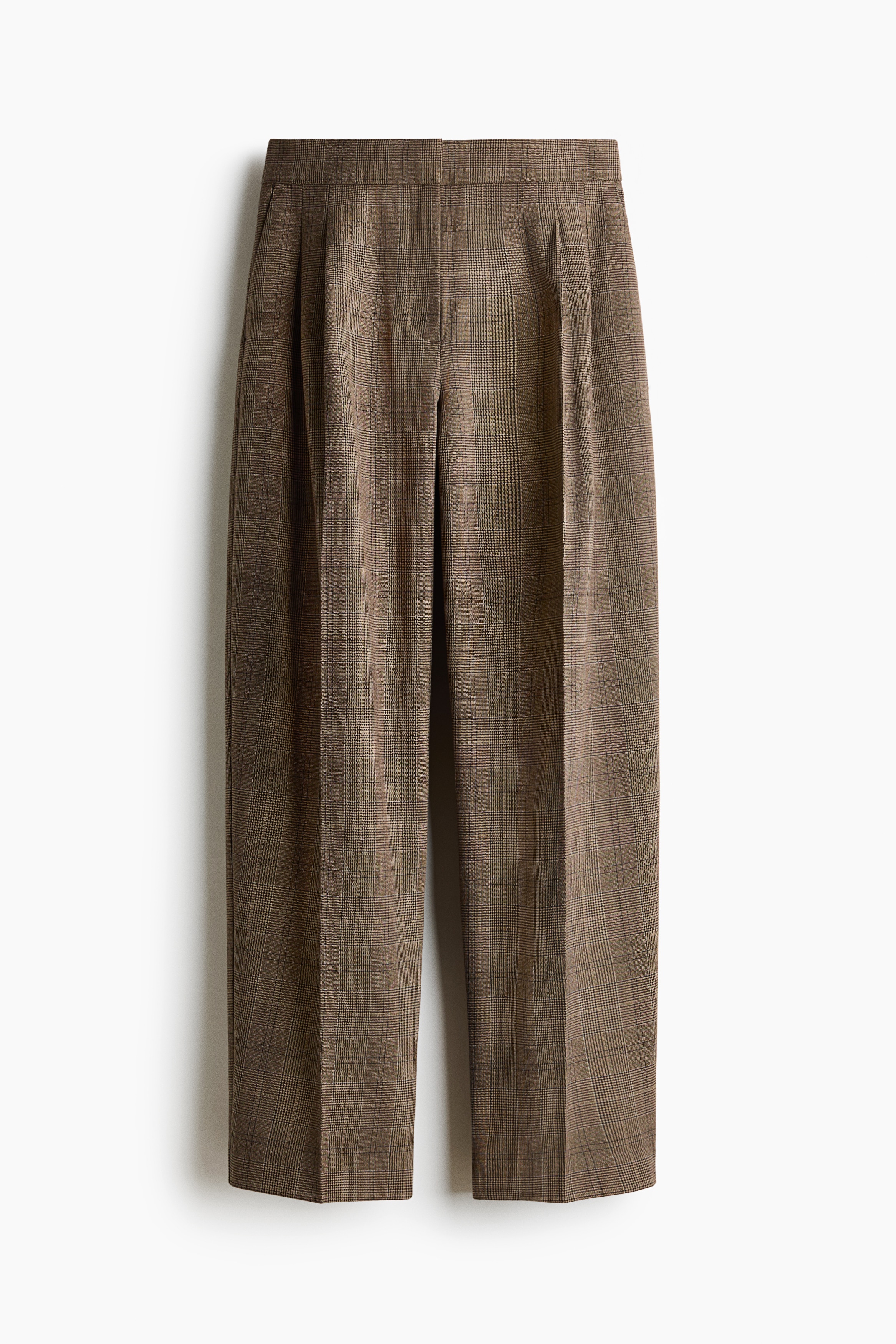 Loose-fit tailored twill trousers in a brown plaid pattern are worn by the model, featuring a high waist, front pleats, and a barrel-leg silhouette with sharp front creases. The trousers are styled with a grey V-neck jumper partially tucked into the waistband and black strappy sandals.
