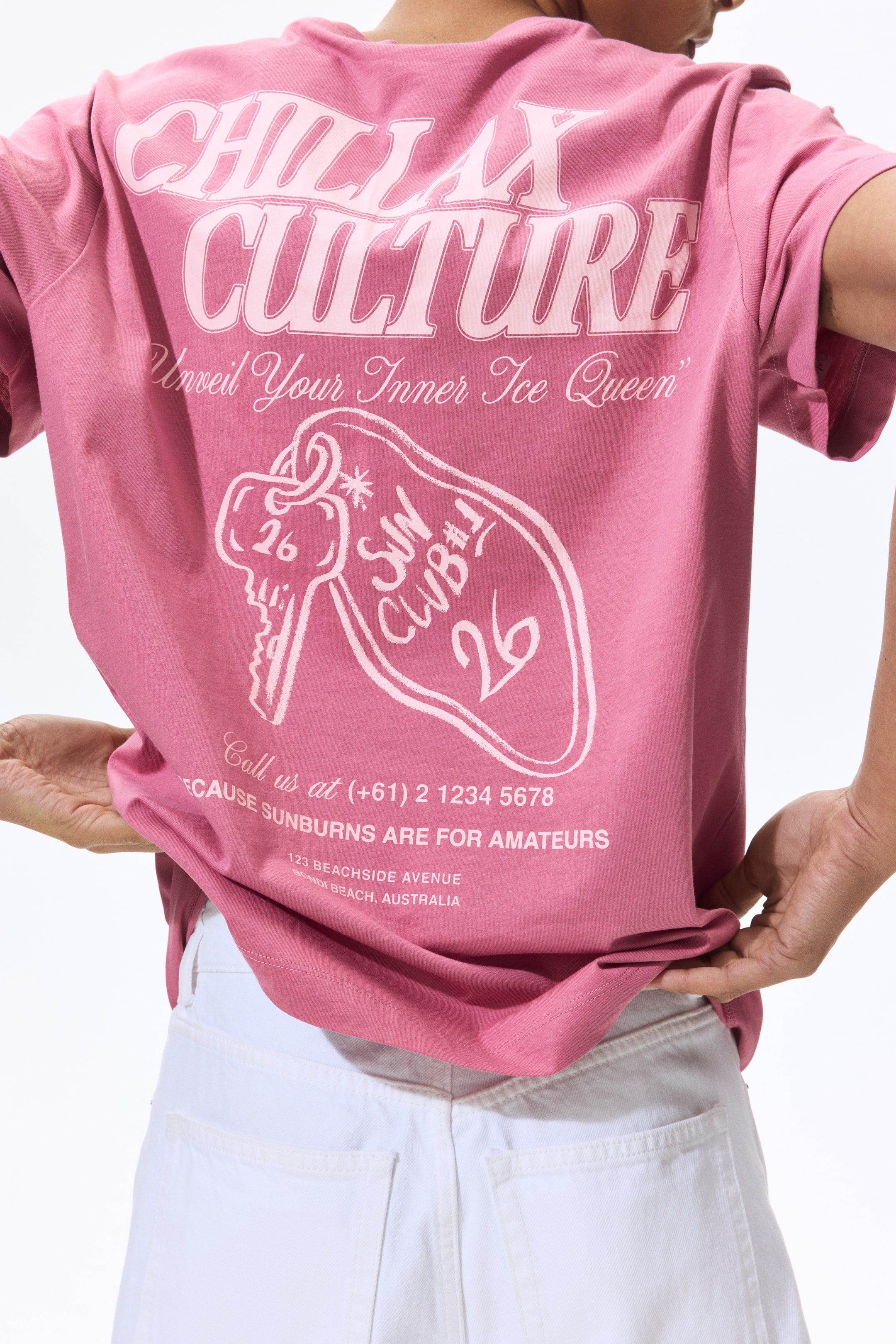 View larger image: A pink short-sleeved t-shirt is shown from the back, showcasing a large light pink graphic print. The design features stylized text that reads "CHILLAX CULTURE" and "Unveil Your Inner Ice Queen," along with a key and a "SUN CLUB #1 26" tag, and additional contact text at the bottom.