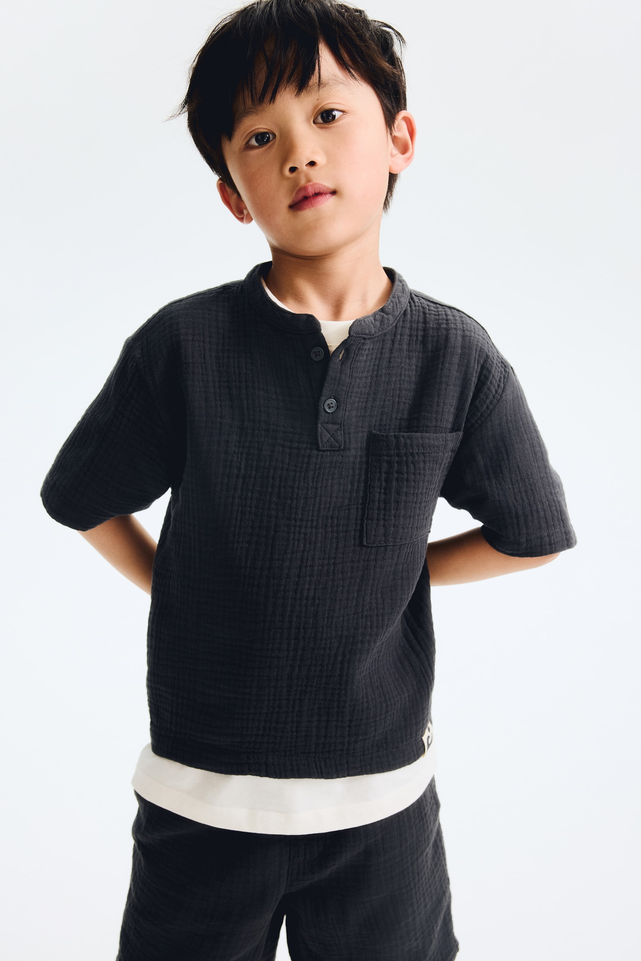 View larger image: This dark-textured, short-sleeve shirt showcases a band collar, a three-button Henley placket, and a left chest patch pocket. It's layered over a white t-shirt, with matching dark-textured shorts visible.
