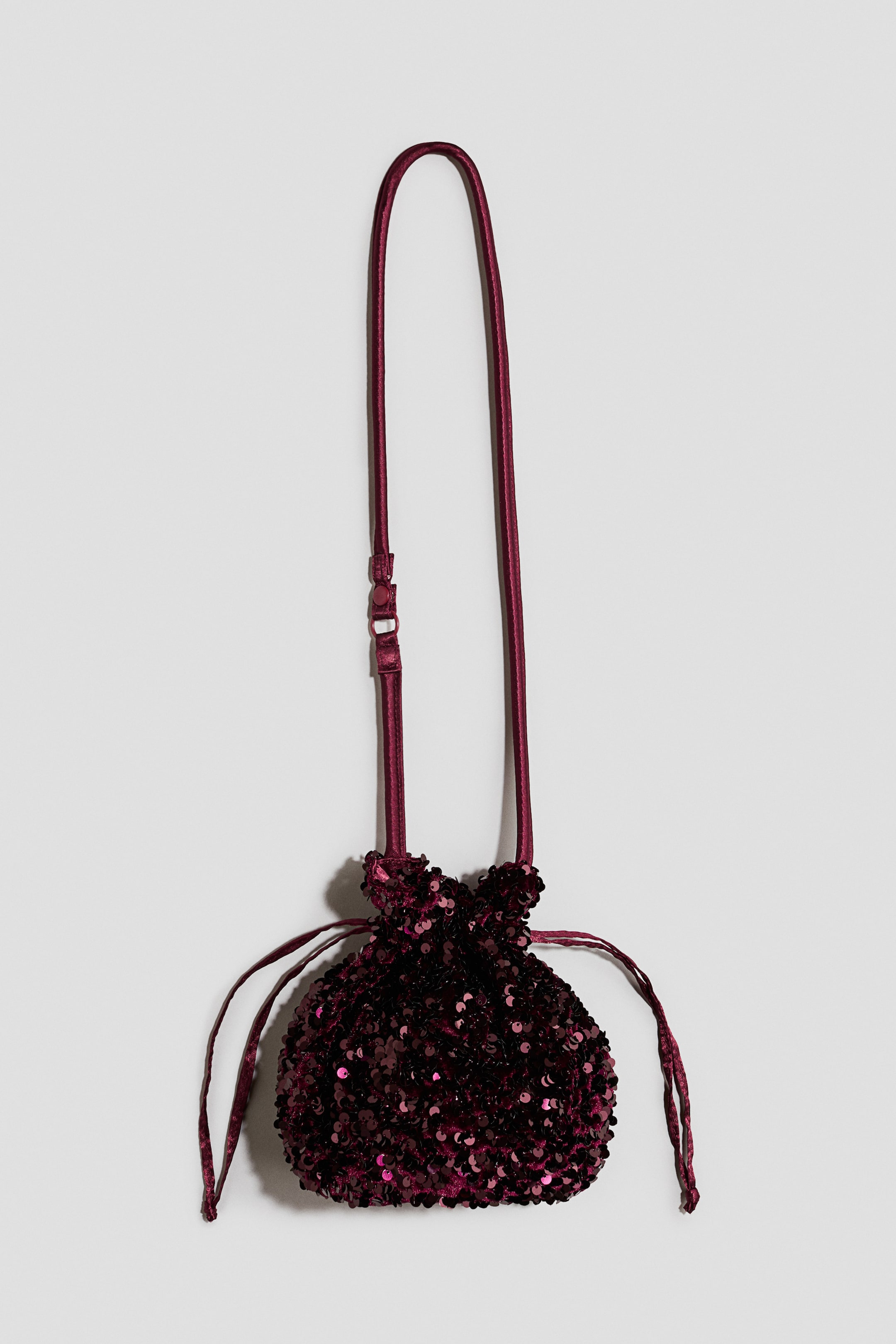 View larger image: A burgundy sequined drawstring bag is hanging from its long, thin satin shoulder strap, facing forwards. The small pouch is covered in shimmering sequins and features a gathered top with thin drawstring ties hanging from the sides.
