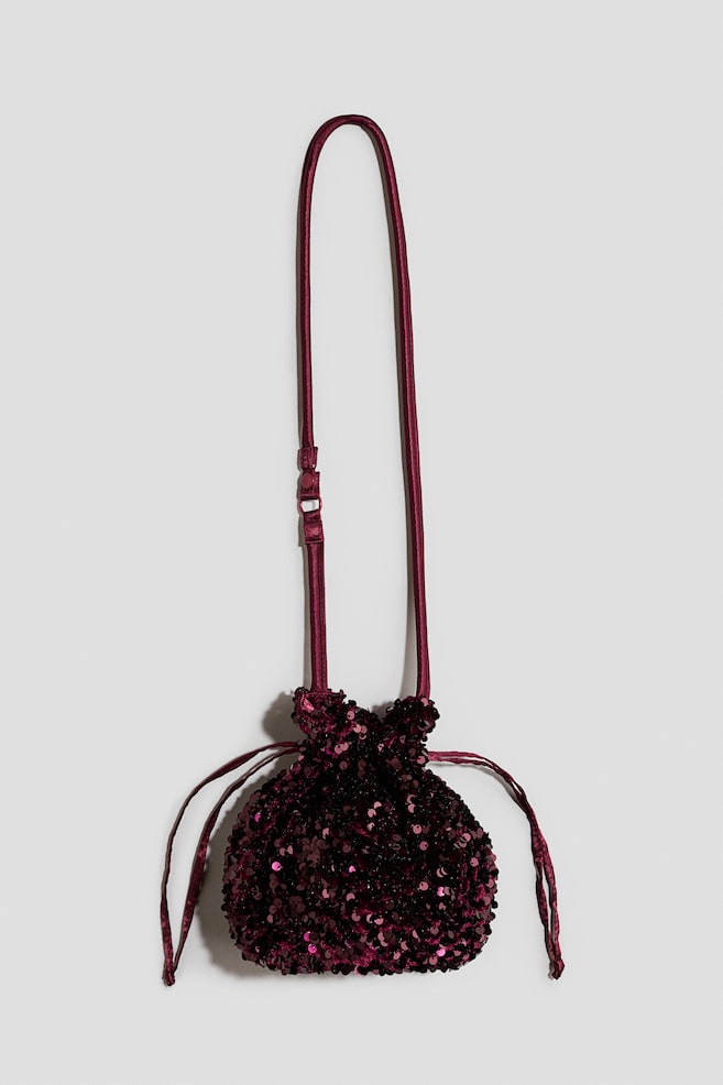 A burgundy sequined drawstring bag is hanging from its long, thin satin shoulder strap, facing forwards. The small pouch is covered in shimmering sequins and features a gathered top with thin drawstring ties hanging from the sides. - 1