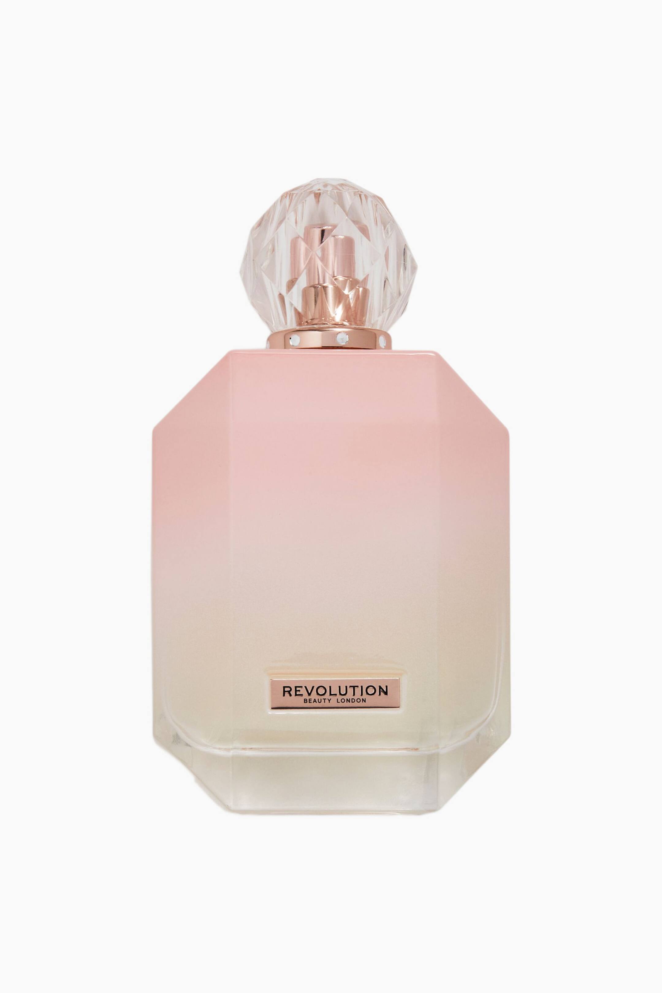 Größeres Bild ansehen: Frontansicht eines Revolution Beauty-Parfümflakons mit facettiertem, achteckigem Glaskörper in sanftem Rosa-zu-Creme-Farbverlauf. Der Flakon zeigt eine klare, diamantgeschliffene geometrische Kappe auf einem Roségold-Kragen mit kleinen eingelassenen Kristallen sowie eine passende Roségold-Plakette nahe der Basis mit der Aufschrift „REVOLUTION BEAUTY LONDON".