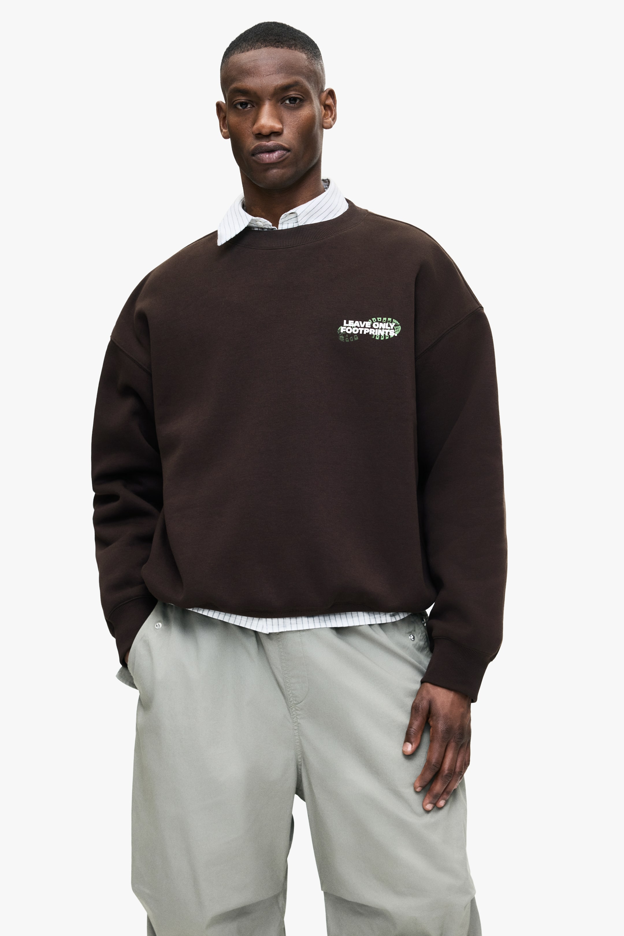 Grösseres Bild ansehen: Ein dunkelbraunes Crewneck-Sweatshirt, das ein grünes und weißes "LEAVE ONLY FOOTPRINTS"-Grafik auf der linken Brust zeigt, wird von dem Model getragen. Darunter wird ein helles Hemd mit Kragen geschichtet, dessen Kragen und Saum sichtbar sind.