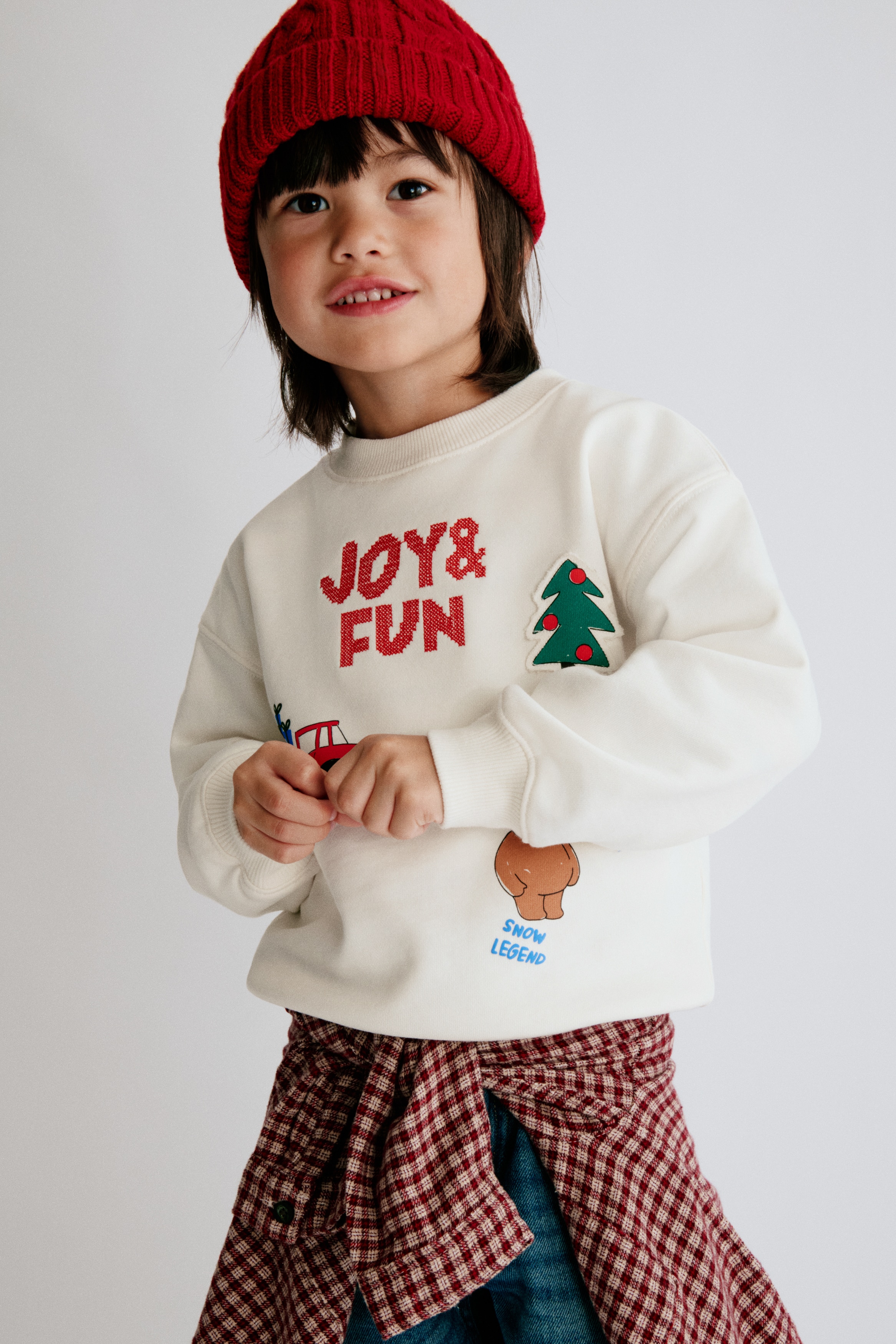 An off-white sweatshirt is shown front-facing, featuring a crew neck, long sleeves with ribbed cuffs, and a ribbed hem. The front features several festive graphics and appliquÃ©s, including the red embroidered text "JOY & FUN", a red toy lorry carrying blue gifts, a green tree patch, and a cartoon reindeer.