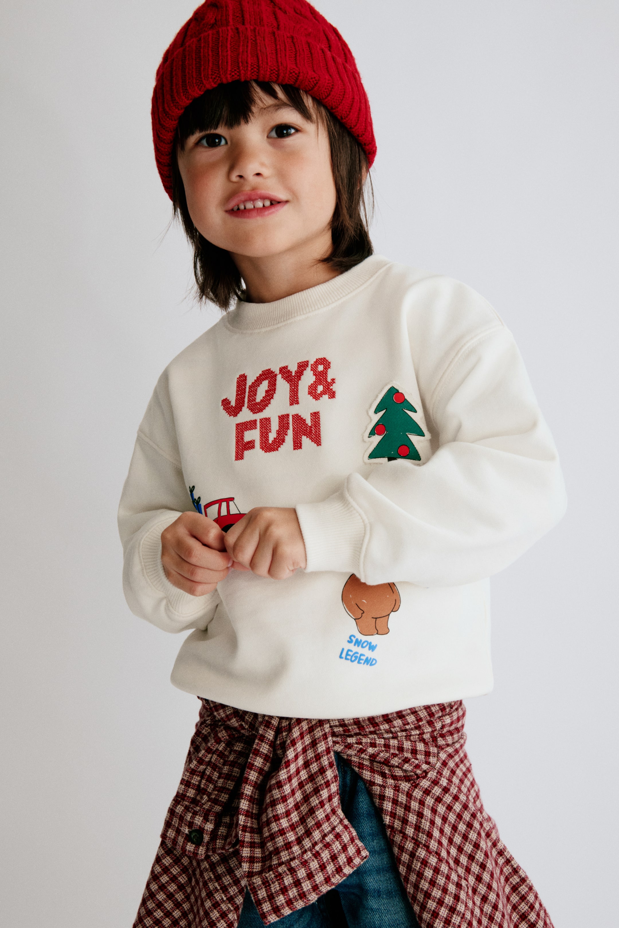 Grotere afbeelding bekijken: Een sweatshirt in losse pasvorm, gebroken wit, wordt van voren getoond en heeft een ronde hals met ribboord, verlaagde schouders en ribgebreide boorden. Op de voorkant staan diverse feestelijke graphics, waaronder de rode tekst 'JOY & FUN', een groen kerstboomembleem en een kleine illustratie van een bruine beer.