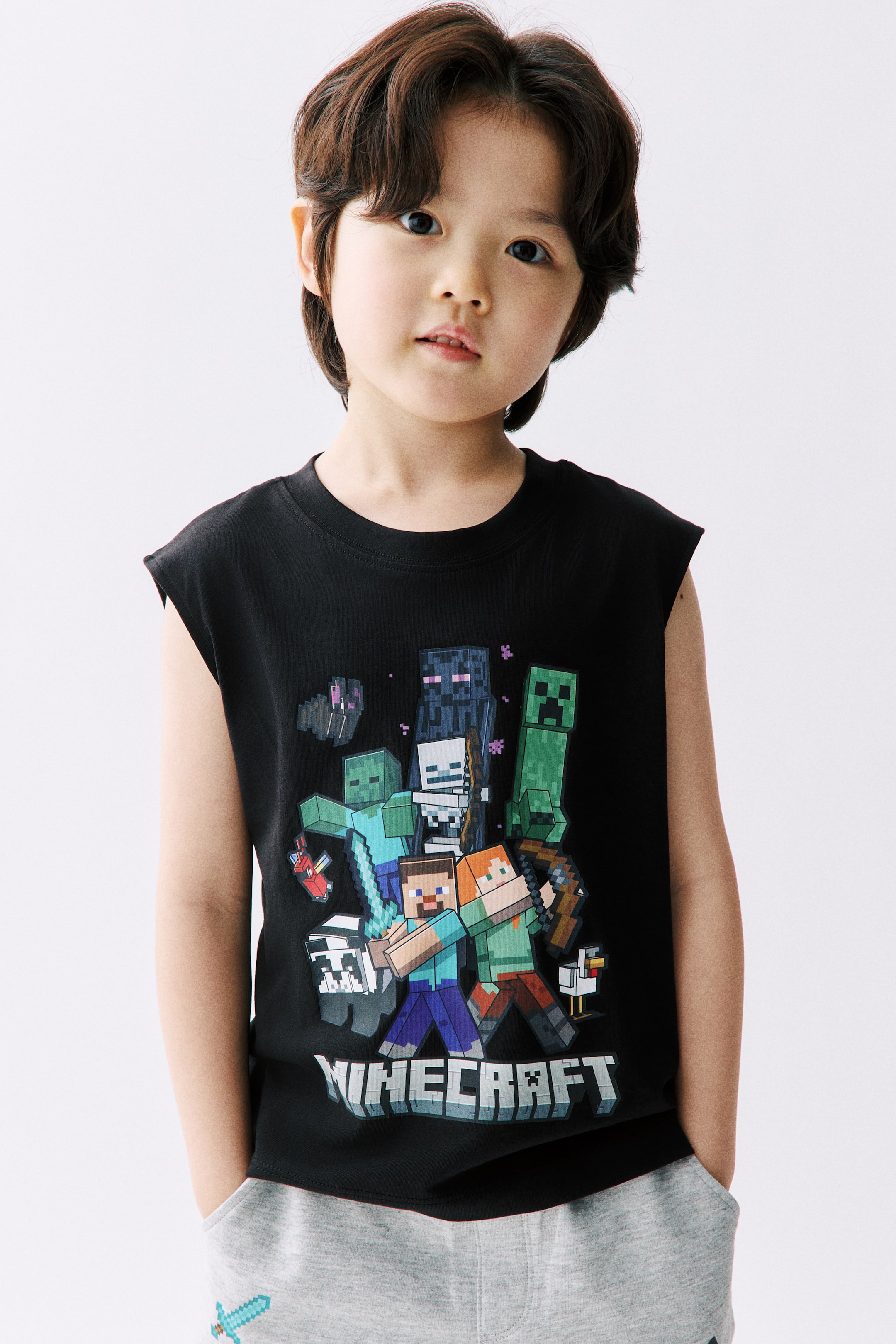 拡大画像を表示: 黒のノースリーブTシャツが正面を向いており、クルーネックと「Minecraft」ロゴを含む様々なMinecraftキャラクターの大きくカラフルなグラフィックプリントが前面に施されています。