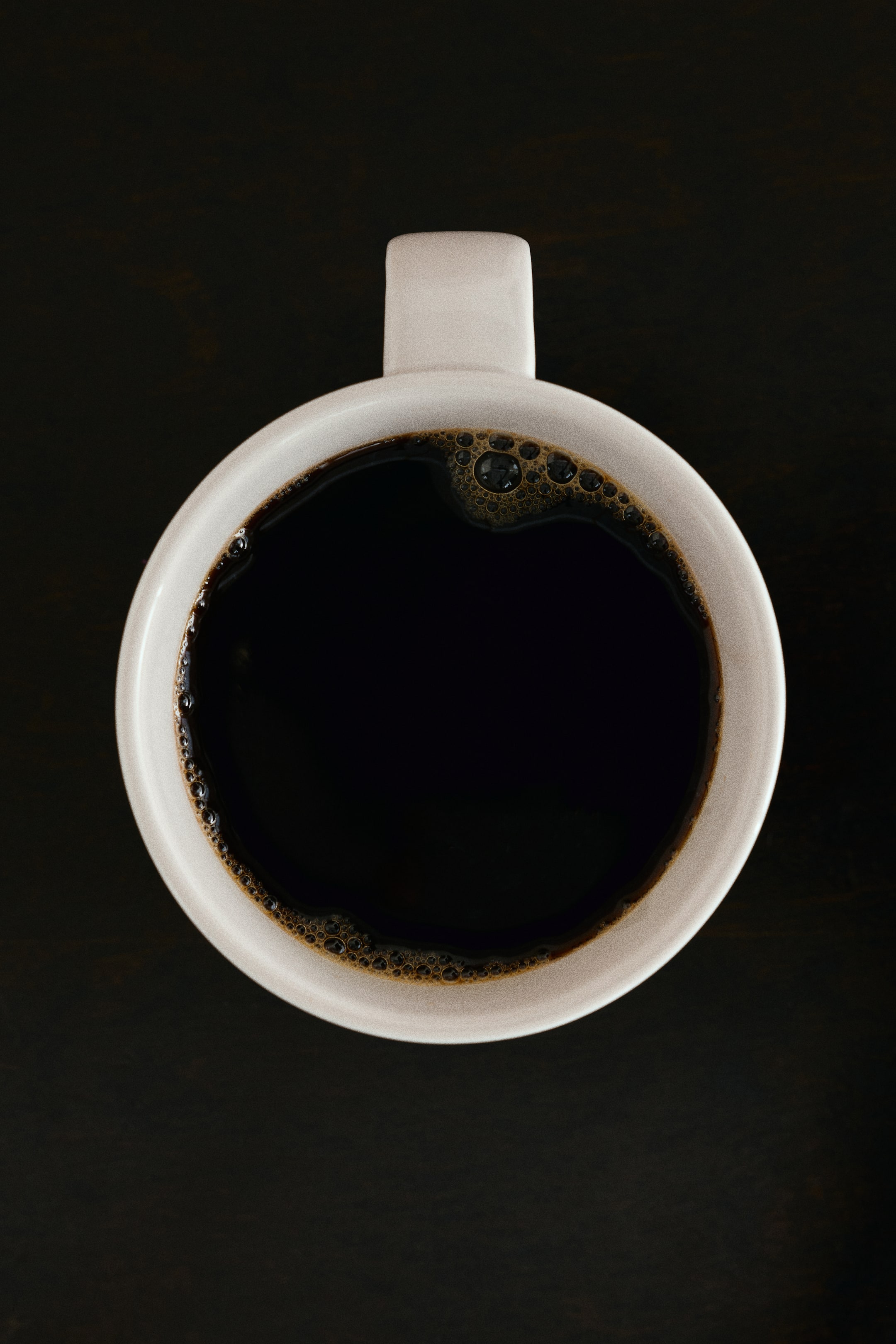 View larger image: A minimalist porcelain mug in a light off-white shade is shown from an overhead perspective, containing black coffee with small bubbles along the inner rim. The mug features a clean, circular shape and a simple rectangular handle.
