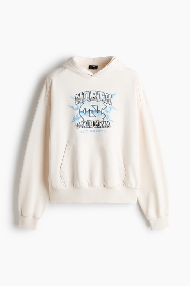Ein cremefarbener Hoodie ist von vorne zu sehen und verfügt über überschnittene Schultern, Rippbündchen, einen gerippten Saum und eine Kängurutasche auf der Vorderseite. Auf der Brust ist ein Grafikdruck mit einem ineinandergreifenden âNCâ-Logo zu sehen, umgeben von dem Schriftzug âNORTH CAROLINA ICE HOCKEYâ sowie hellblauen Blitzdetails. - 1