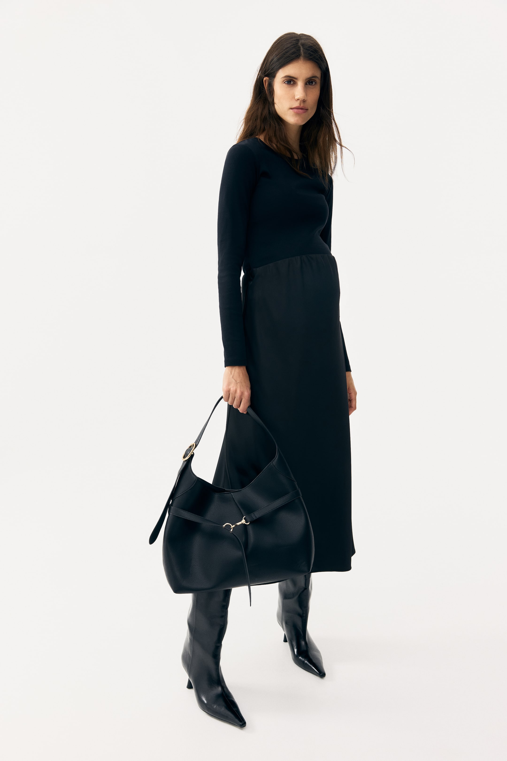 Visualizza immagine più grande: La borsa hobo in pelle nera martellata è tenuta, leggermente inclinata in avanti e verso sinistra, mostrando la sua morbida forma a mezzaluna. Presenta un unico manico con fibbia dorata e una cinghia decorativa anteriore con ferramenta a morsetto dorato, da cui pende un sottile tirante in pelle.