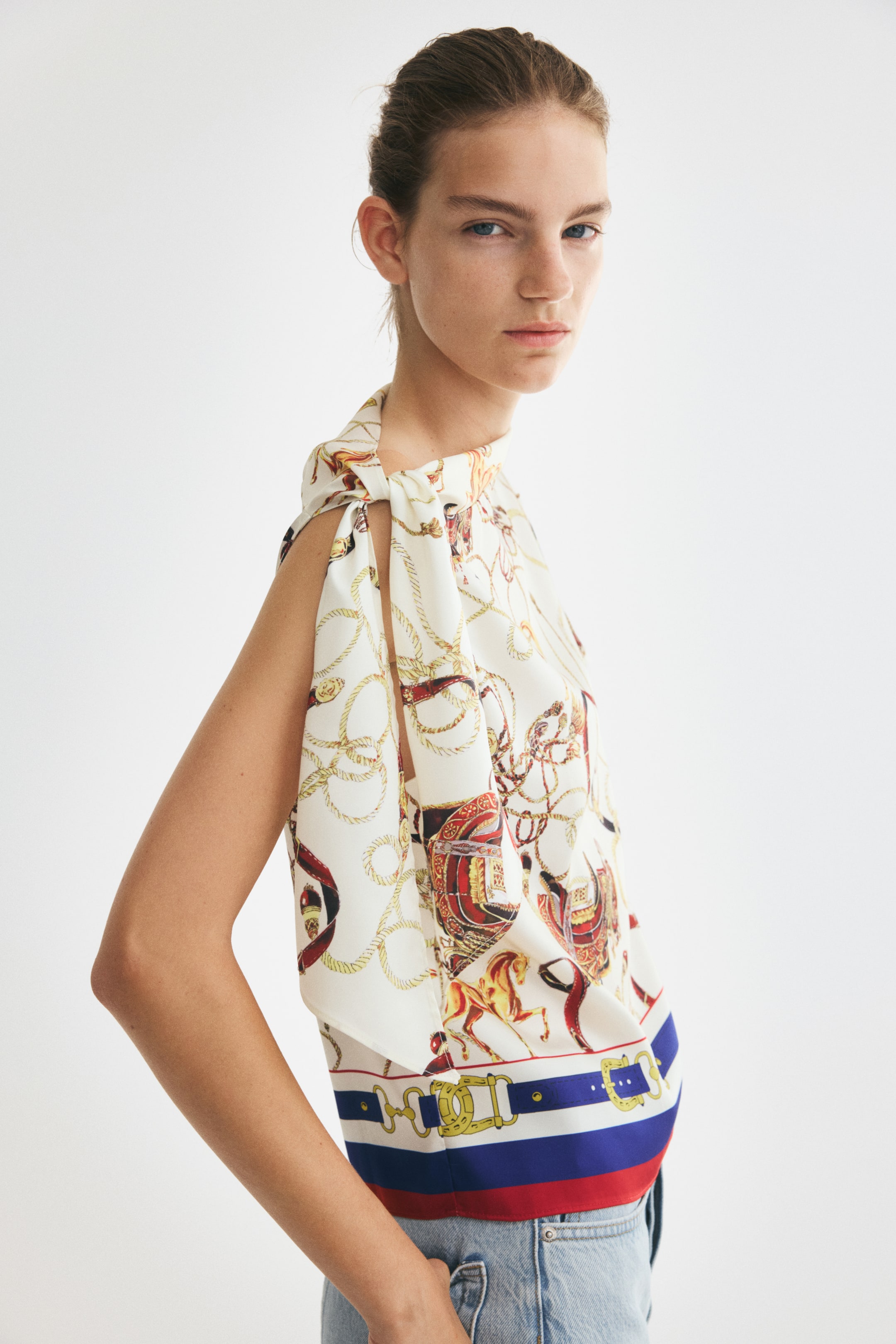 Grotere afbeelding bekijken: Een top van satijnkeper met patroon en een print met een ruiterthema in rood, goud en blauw wordt gedragen door het model. De top heeft een asymmetrische halslijn met een brede strik die over één schouder valt en is ingestopt in lichtgewassen denim jeans.