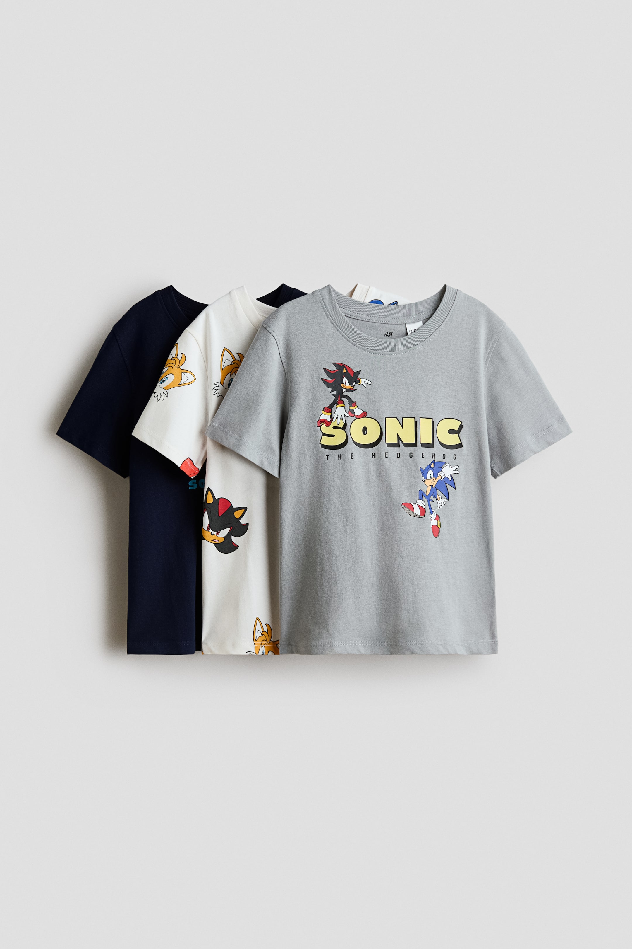 Grotere afbeelding bekijken: Drie klassieke T-shirts van zachte katoenjersey liggen overlappend en zijn van voren gericht om hun designs en ribgebreide ronde halslijnen te tonen. Het grijze T-shirt op de voorgrond heeft een centrale grafische print van Sonic the Hedgehog, terwijl het gebroken witte T-shirt in het midden een all-over karakterprint laat zien en het marineblauwe T-shirt achteraan gedeeltelijk zichtbaar is.