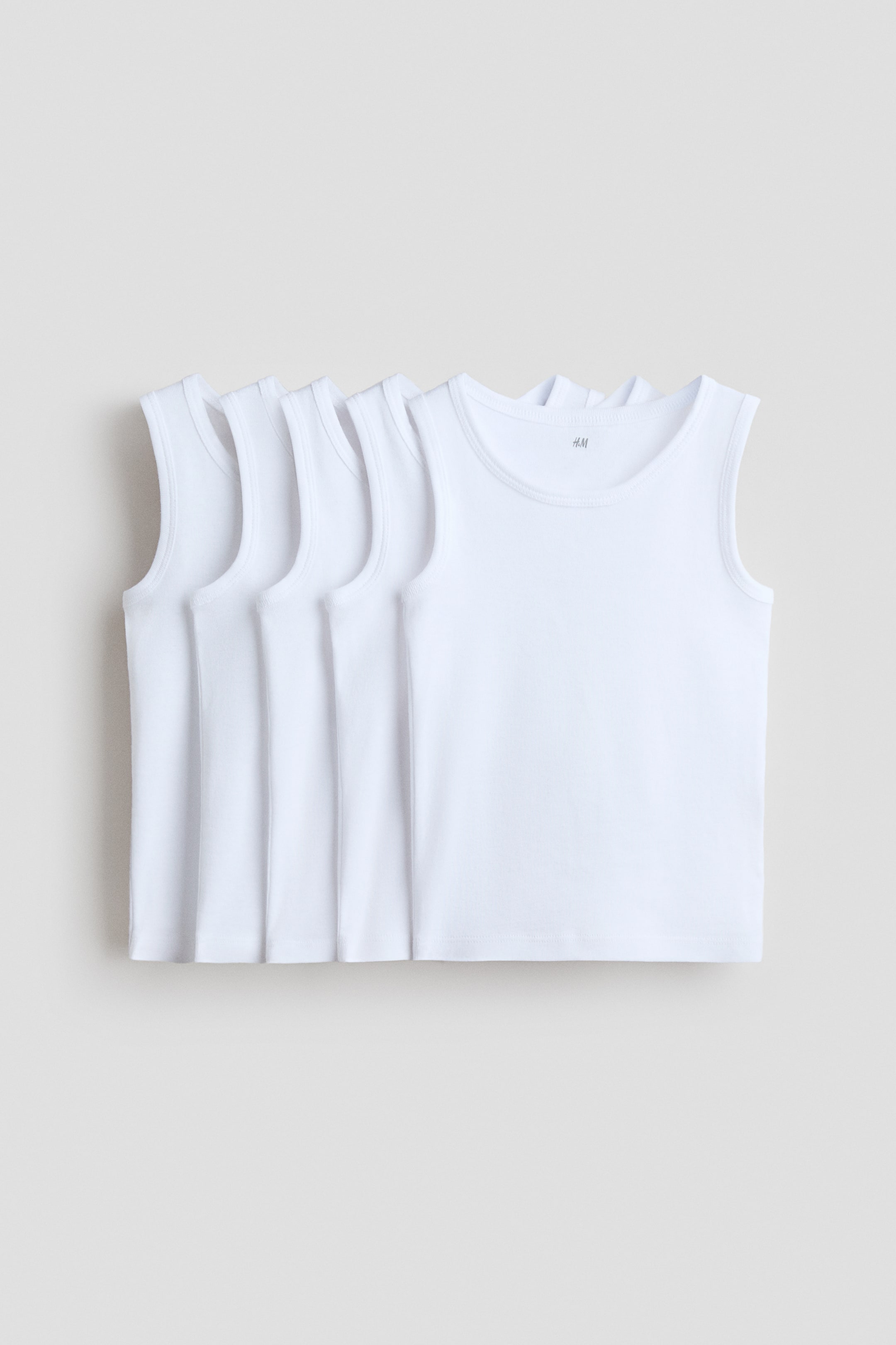 View larger image: Five white tank tops are displayed in a layered manner, with the front tank top facing forward. Each tank top showcases a round neck and ribbed trim along the neck and armholes.