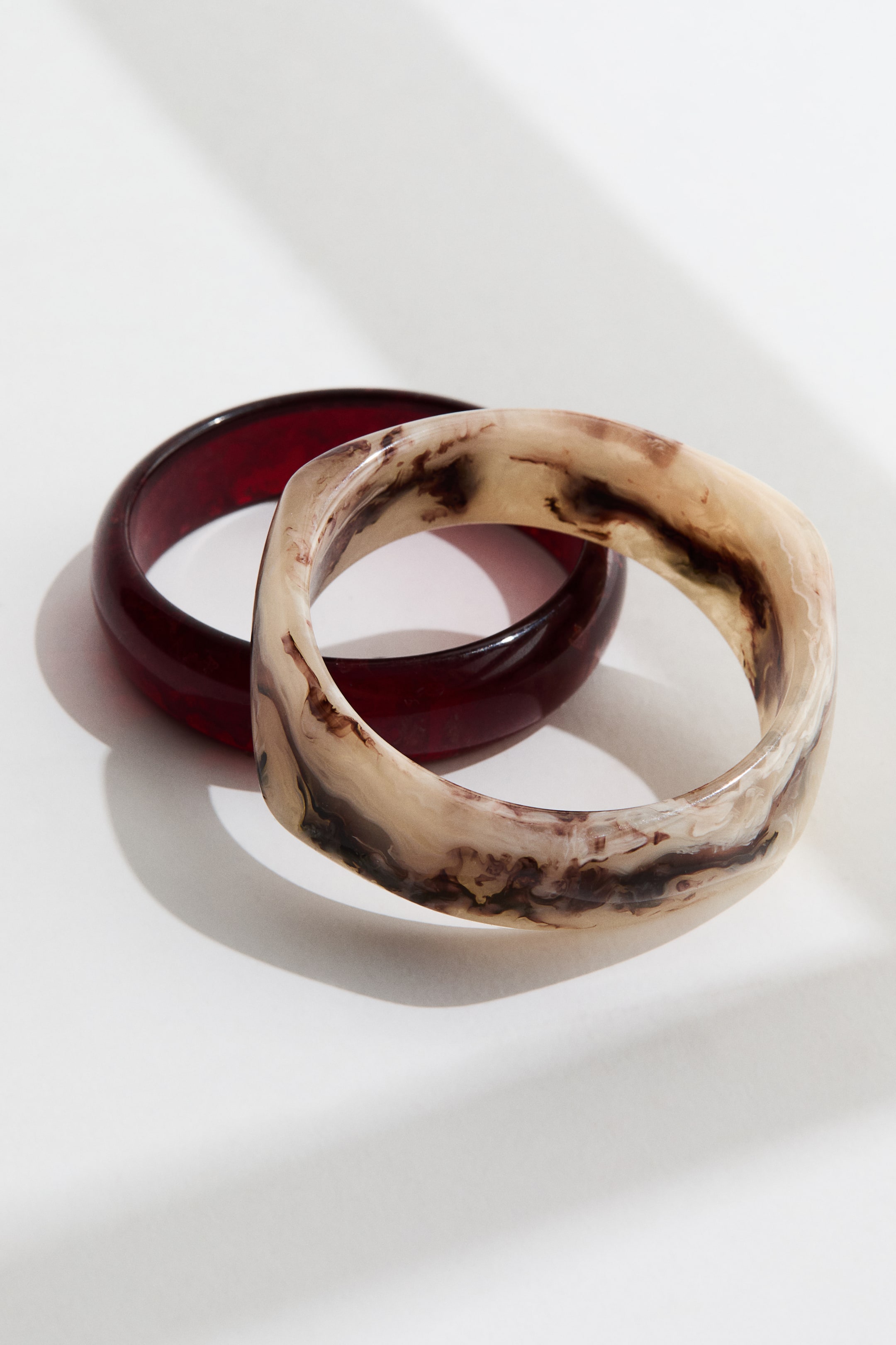 View larger image: The marbled beige, brown, and off-white bangle with a faceted, slightly squared shape is overlapping a round, translucent deep red bangle.