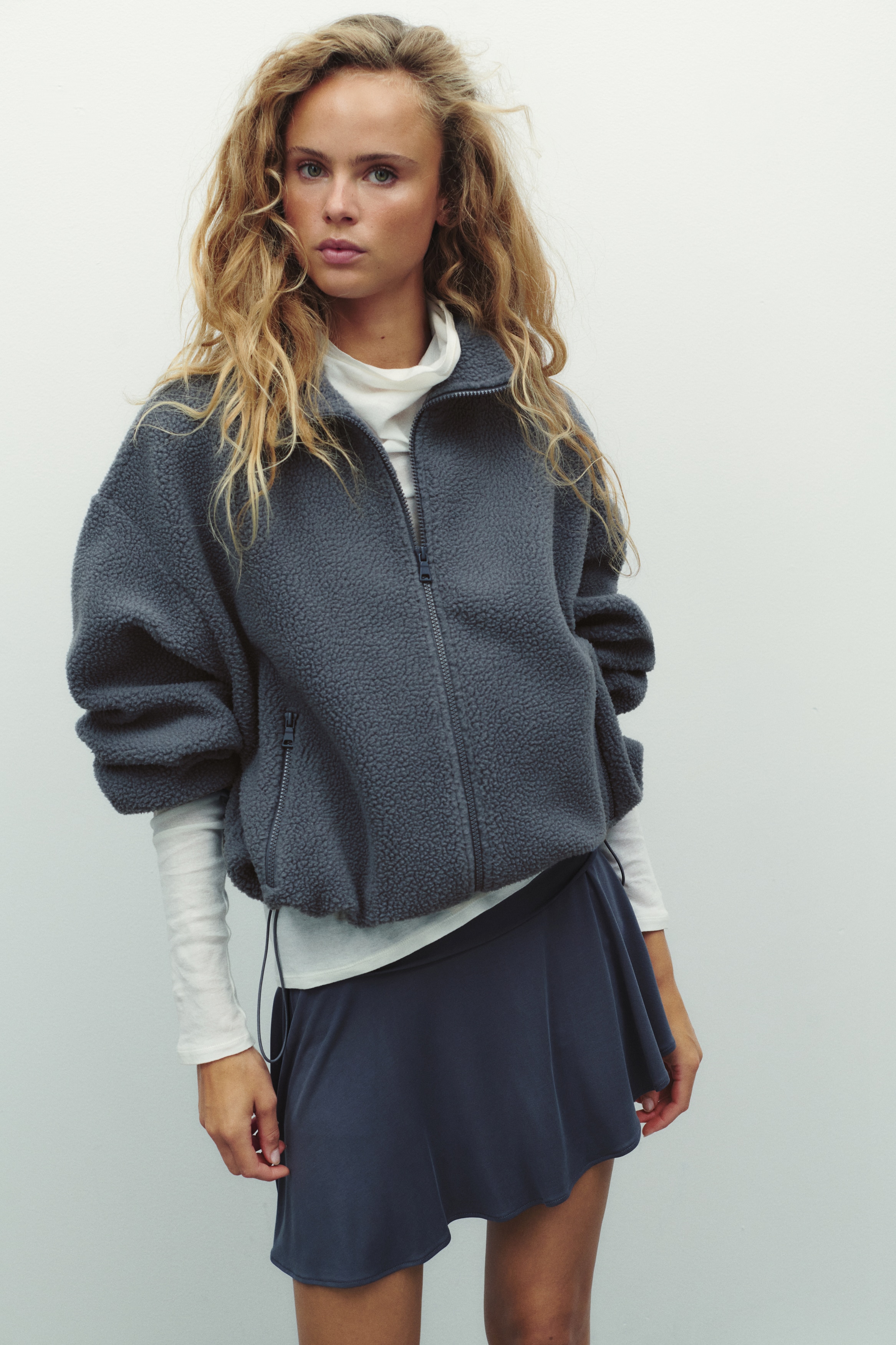 A dark grey teddy fleece jacket is shown from the front, featuring a full-length zip fastening and a stand collar. It has two zipped pockets on the front, with long sleeves finished with elasticated cuffs.