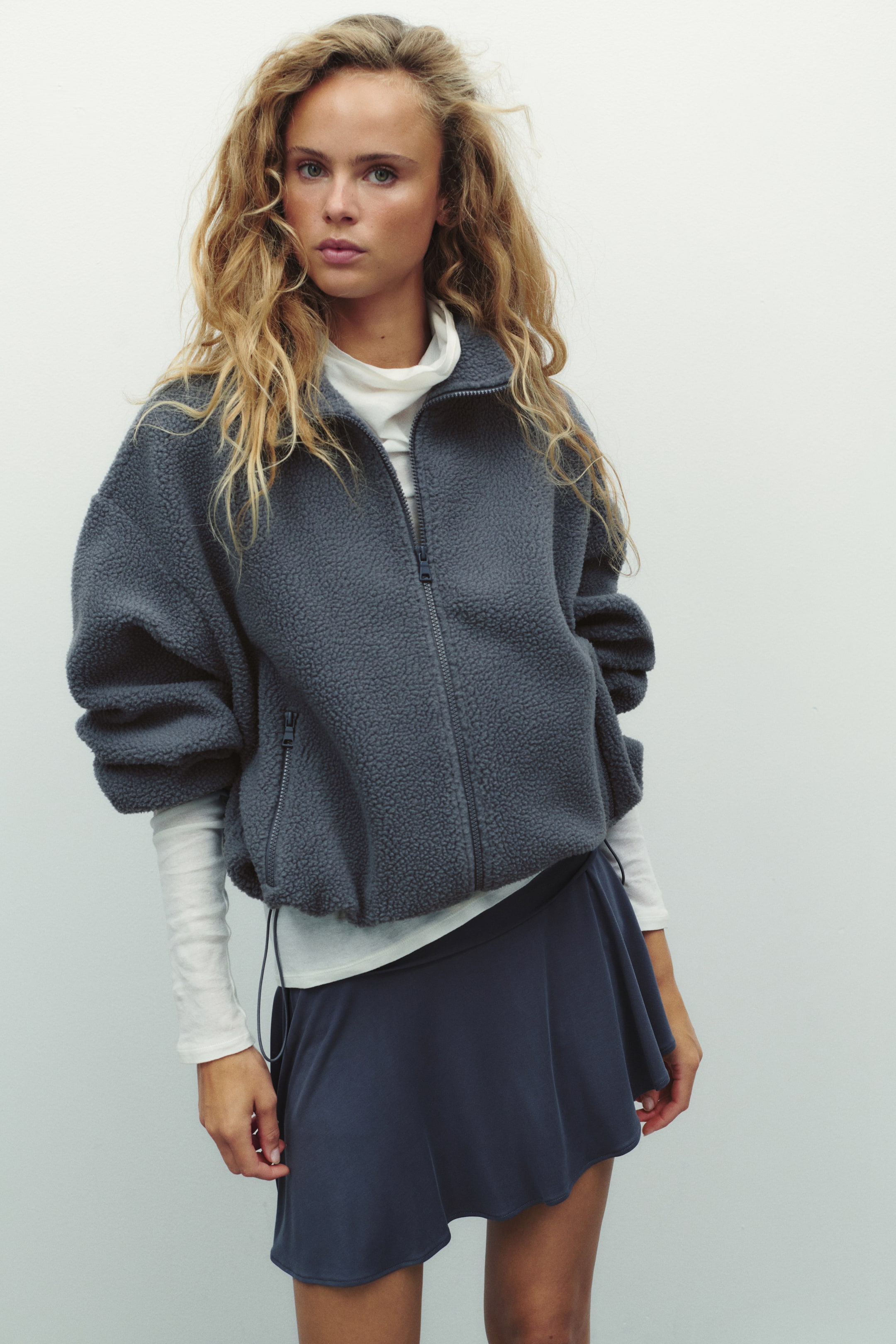 View larger image: The model is wearing a loose-fit grey teddy jacket with a stand collar, front zip closure, and zipped side pockets. The jacket is layered over a white long-sleeved high-neck top and paired with a dark blue skirt.