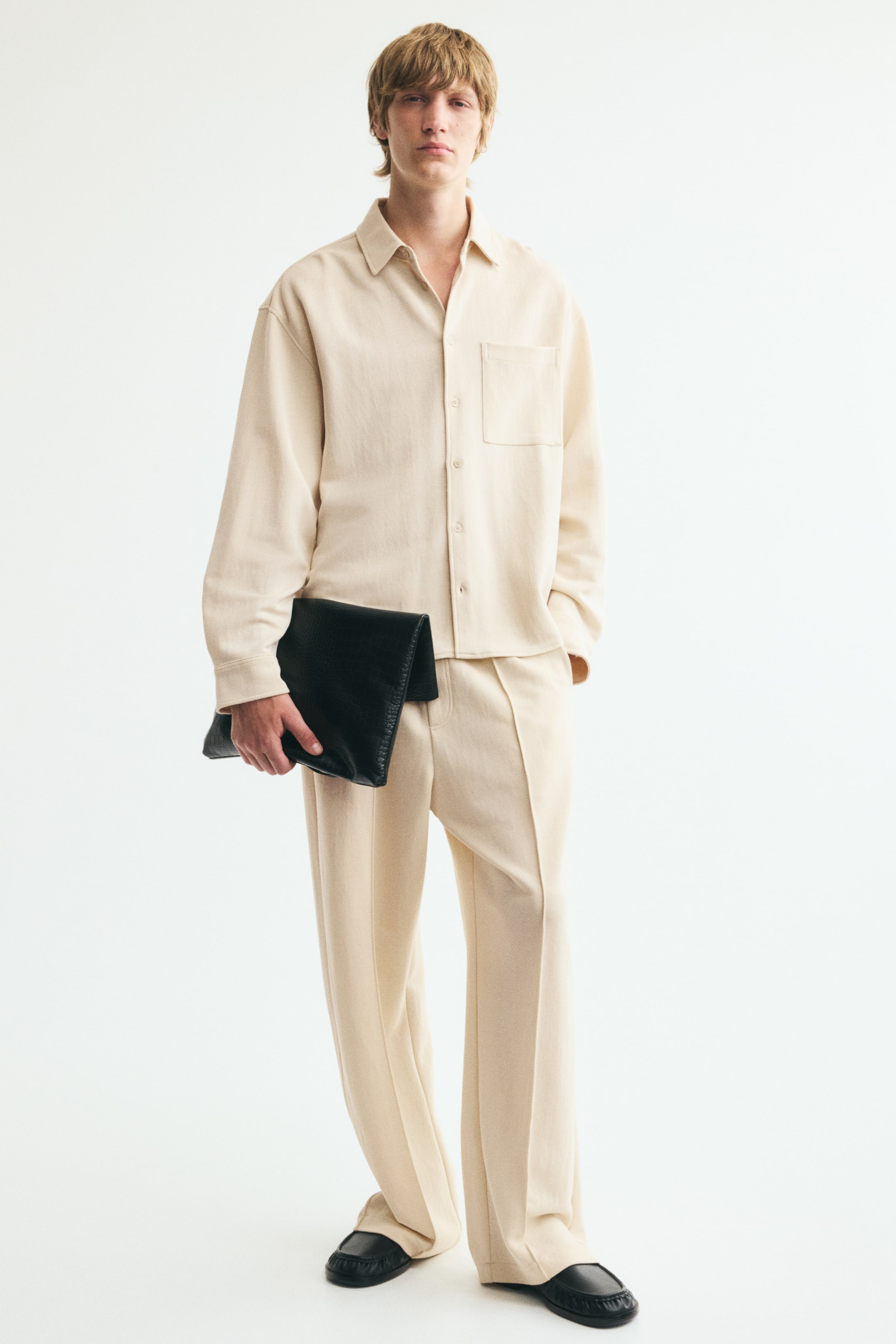 View larger image: The model is wearing joggers in interlock jersey, featuring a relaxed fit, a faux fly, side pockets, and front creases. The joggers are styled with a cream shirt and black shoes.