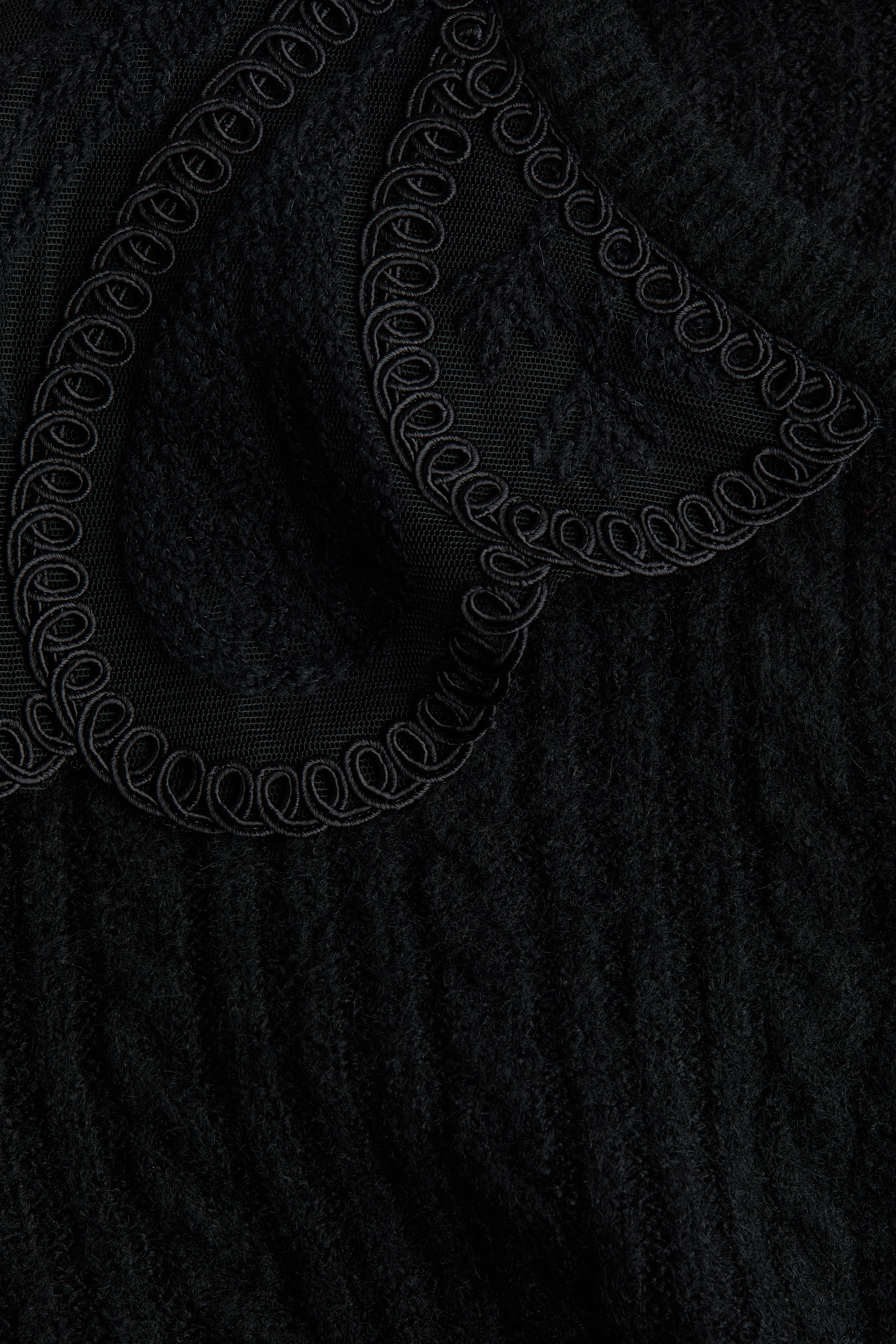 拡大画像を表示: 黒いカーディガンは、柔らかいケーブルニットの生地とレースの襟のクローズアップを特徴としています。襟には、渦巻くようなループ状の模様が入った複雑な刺繍の縁取りが施されています。